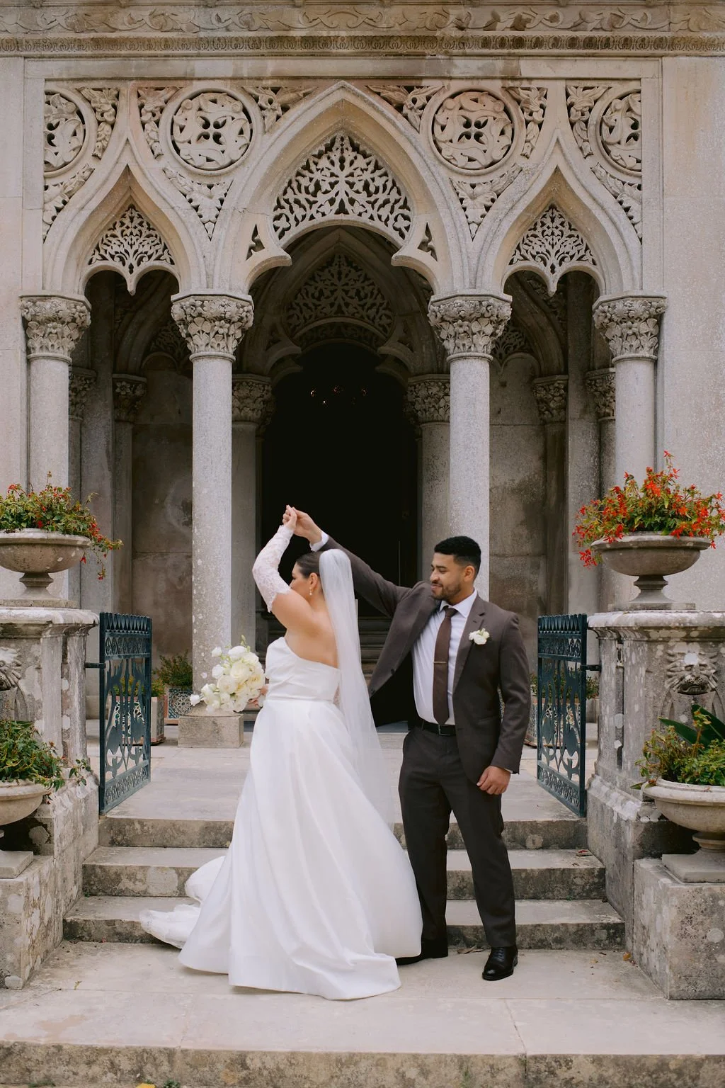 A bride and groom dancing outside a historic stone building with ornate arches and columns, surrounded by flower arrangements.