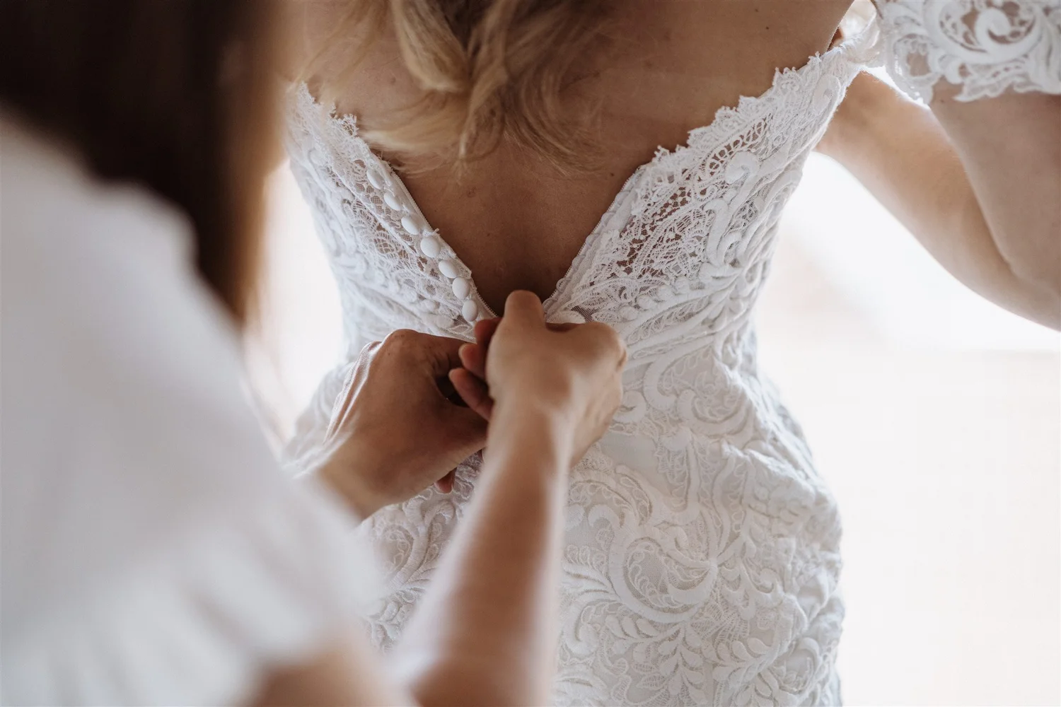 A person wearing a white lace wedding dress is being buttoned up at the front by another person.