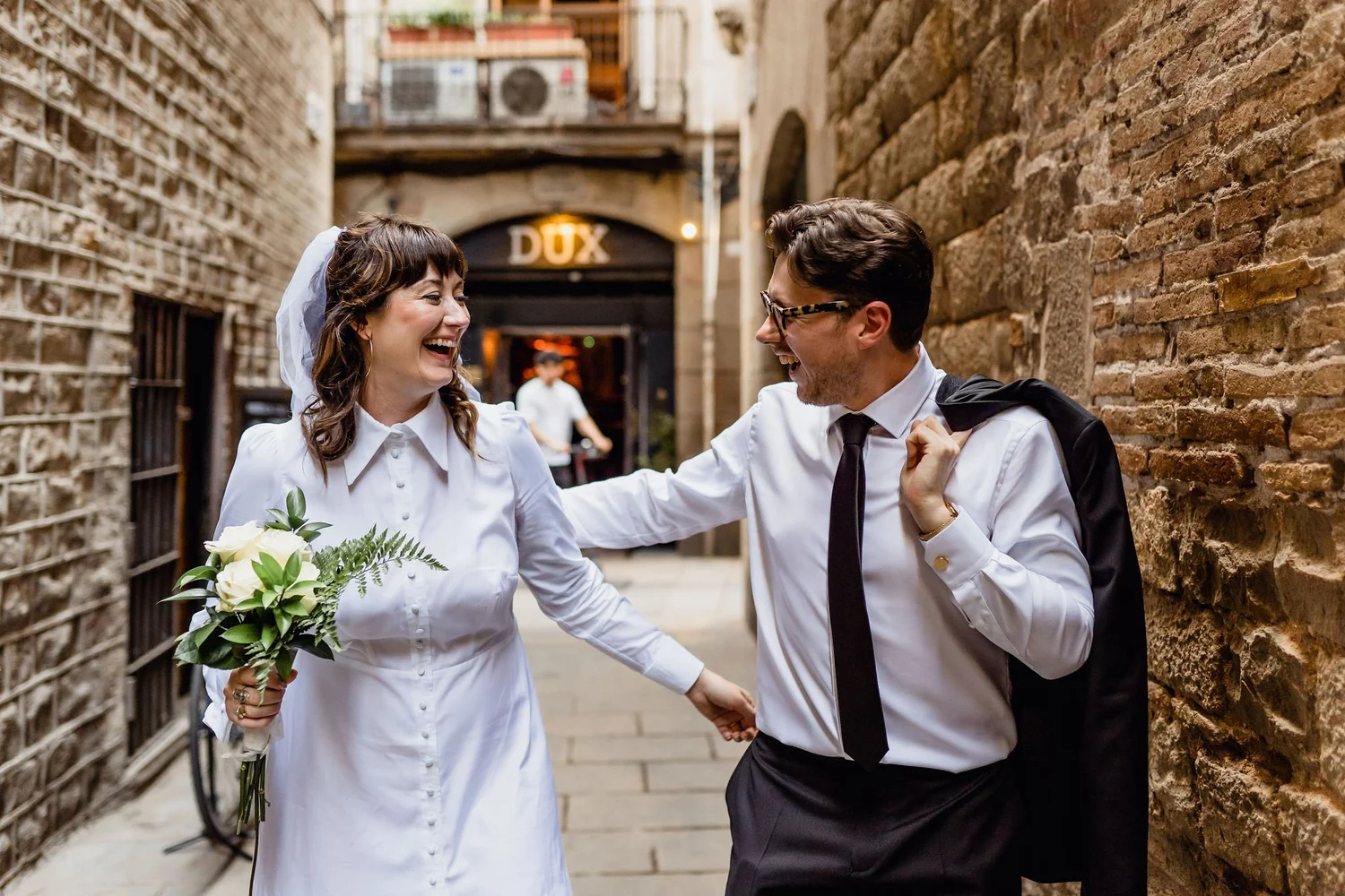 A woman in a white shirt holding a bouquet of white flowers and a man in a white shirt and tie, smiling and holding hands in a narrow alley with brick walls, with a sign that reads 'DUX' in the background.
