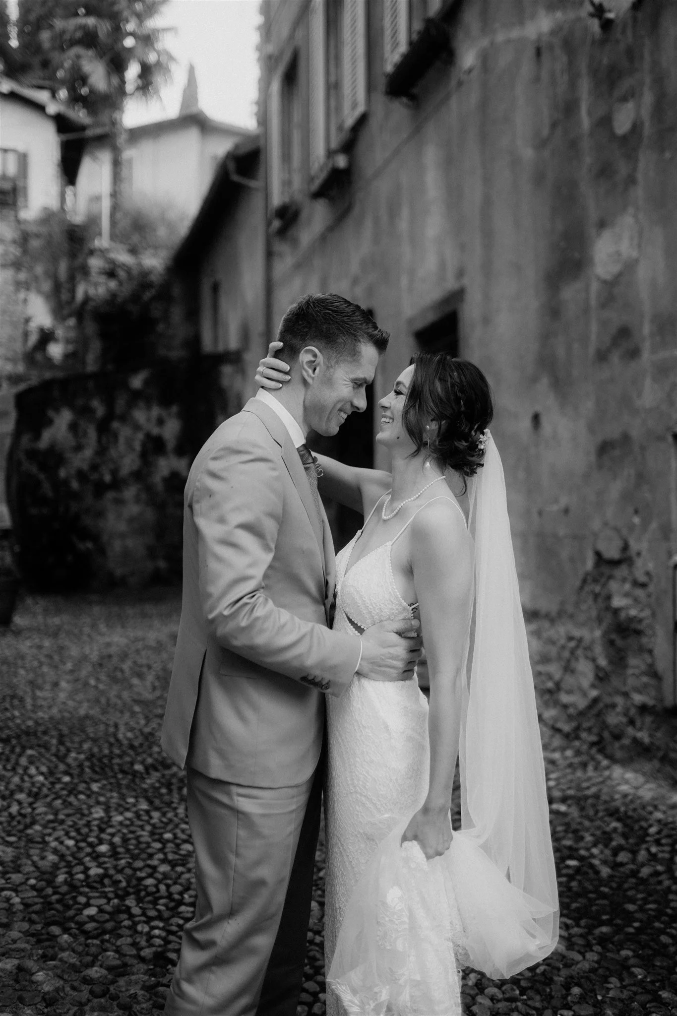 A bride and groom share an intimate moment outdoors, smiling and holding each other closely, with a stone building and trees in the background.
