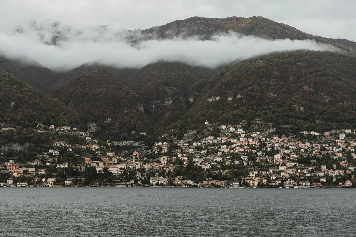 A coastal town at the base of green, forested mountains shrouded in clouds, with buildings along the shoreline and a body of water in the foreground.