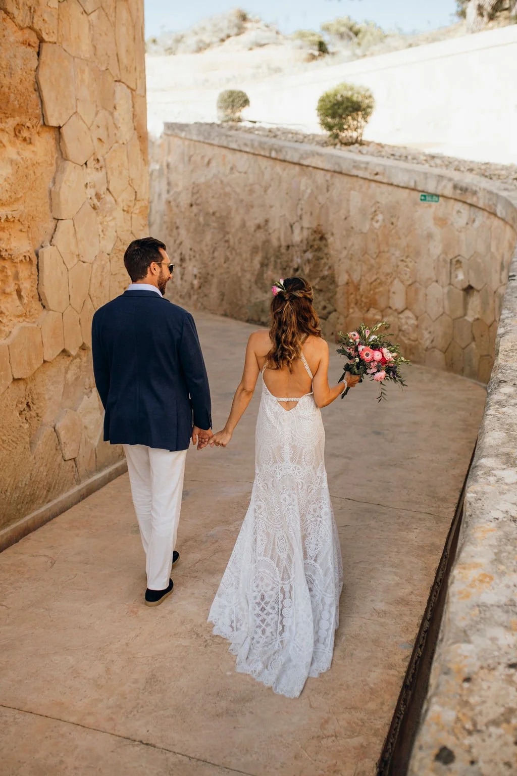 A couple, a man in a navy blazer and white pants and a woman in a white lace wedding dress, hold hands and walk together, with the woman holding a bouquet of pink flowers, along a stone pathway with a stone wall on one side and open outdoor scenery i