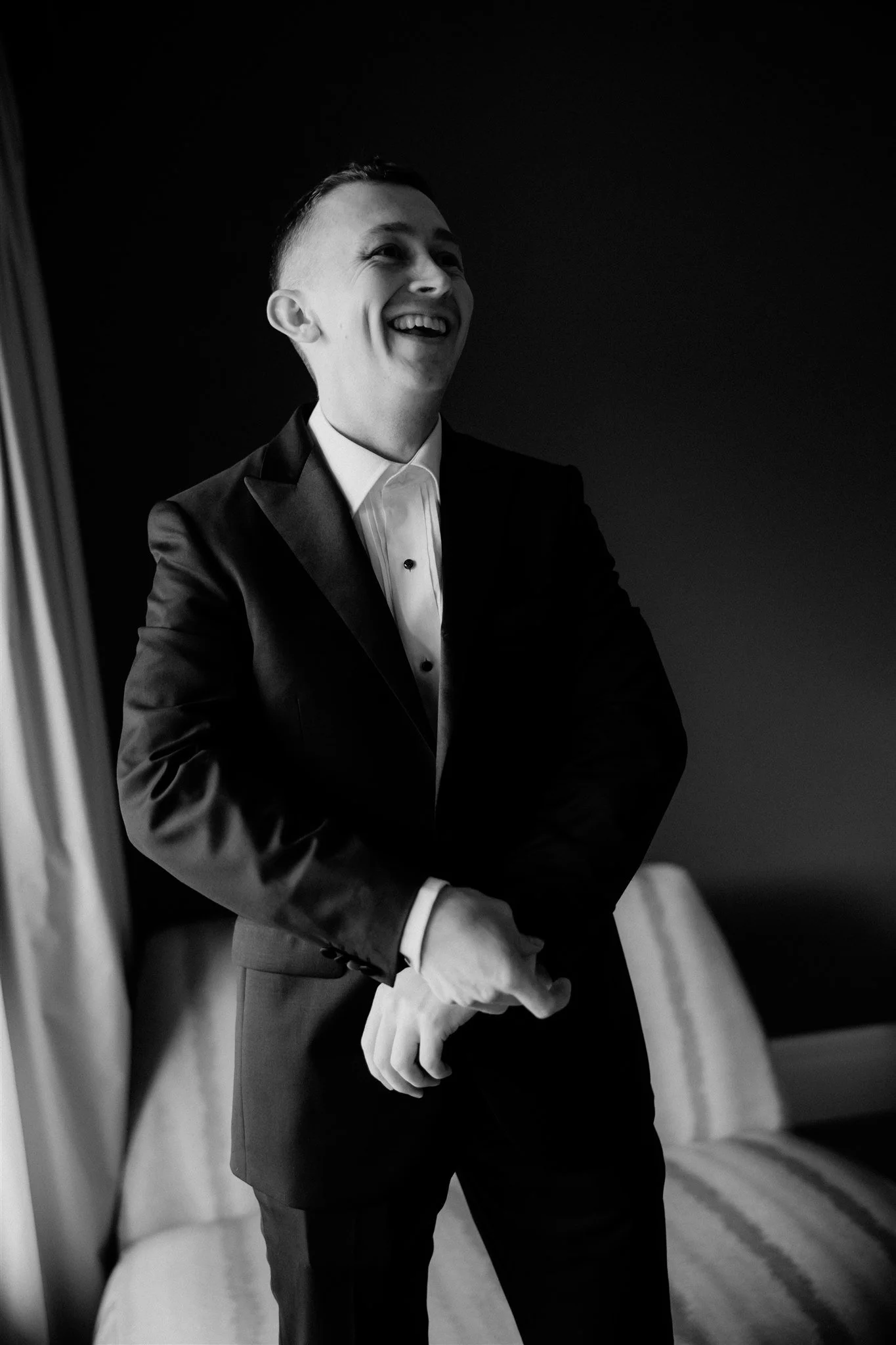 A black and white photo of a man in a tuxedo smiling against a dark background.