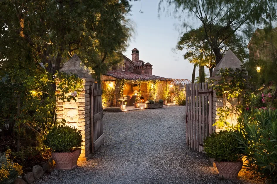 An outdoor garden area at dusk with an open wooden gate, potted plants, and a gravel pathway leading to a cozy, well-lit patio with chairs and a small table.