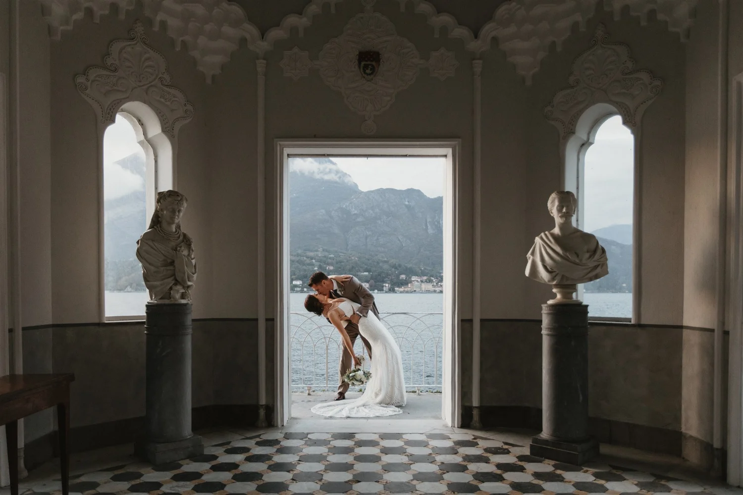 A newlywed couple sharing a kiss and dip on a balcony with lake and mountain view, inside a historic building with ornate interior and sculptures.