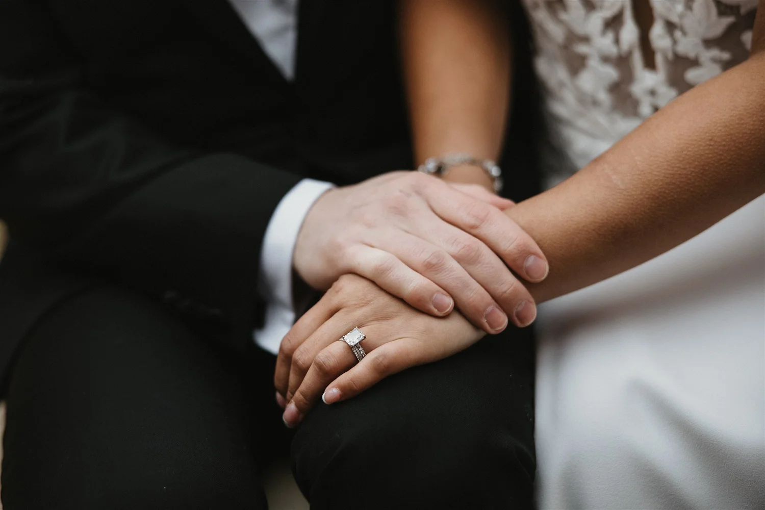 A couple holding hands, with the focus on the woman's hand showing a wedding ring, and the man's hand holding hers. The woman is wearing a diamond bracelet and a floral dress.
