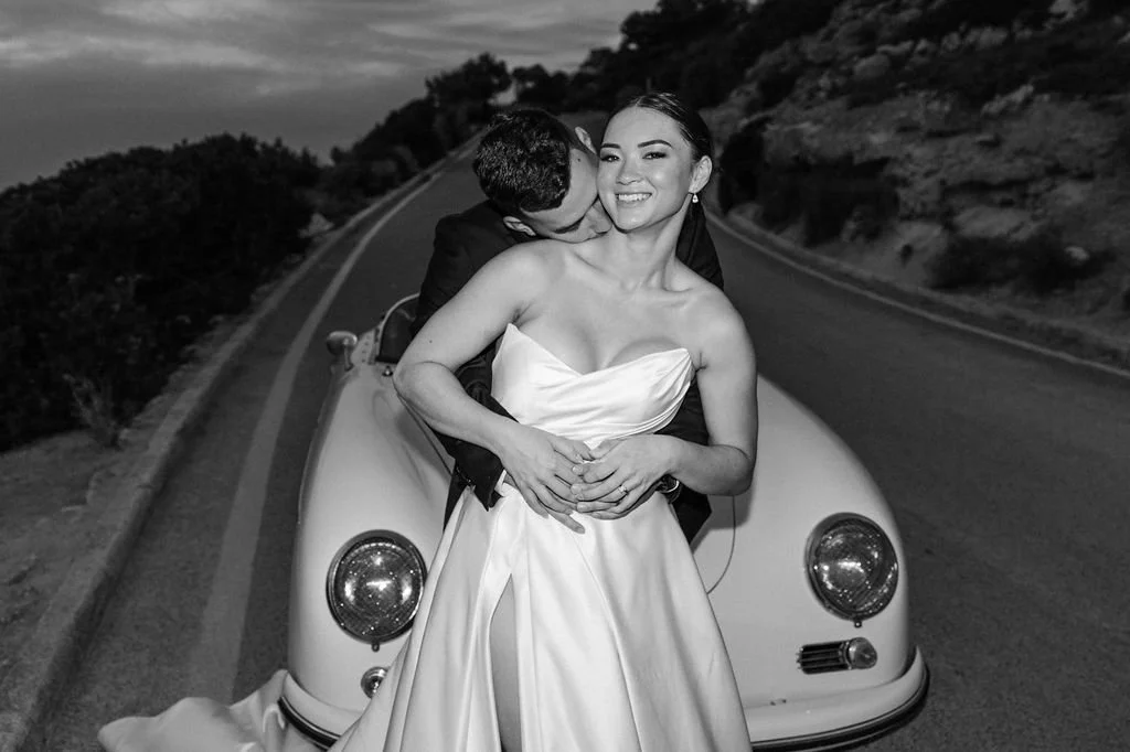 A smiling woman in a bridal gown and a man in a suit posing together in front of a vintage car on a road with hills in the background, black and white photo.