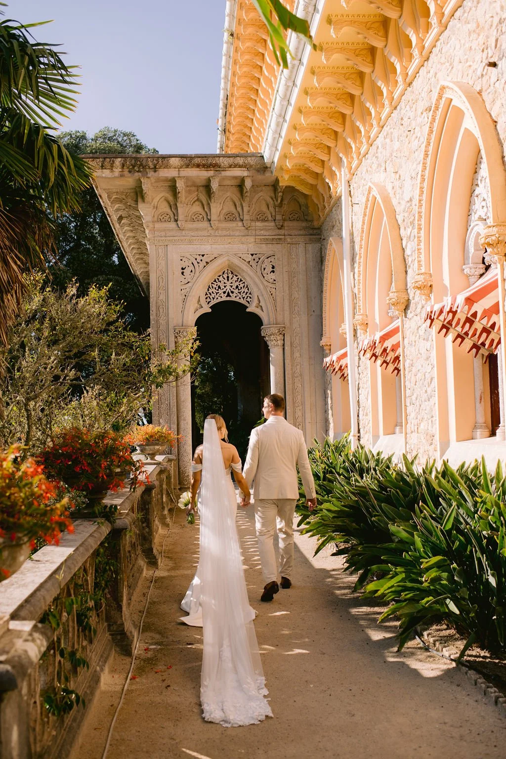 A bride and groom walking hand-in-hand outside a historic building with ornate architecture, lush greenery, and colorful flowers.
