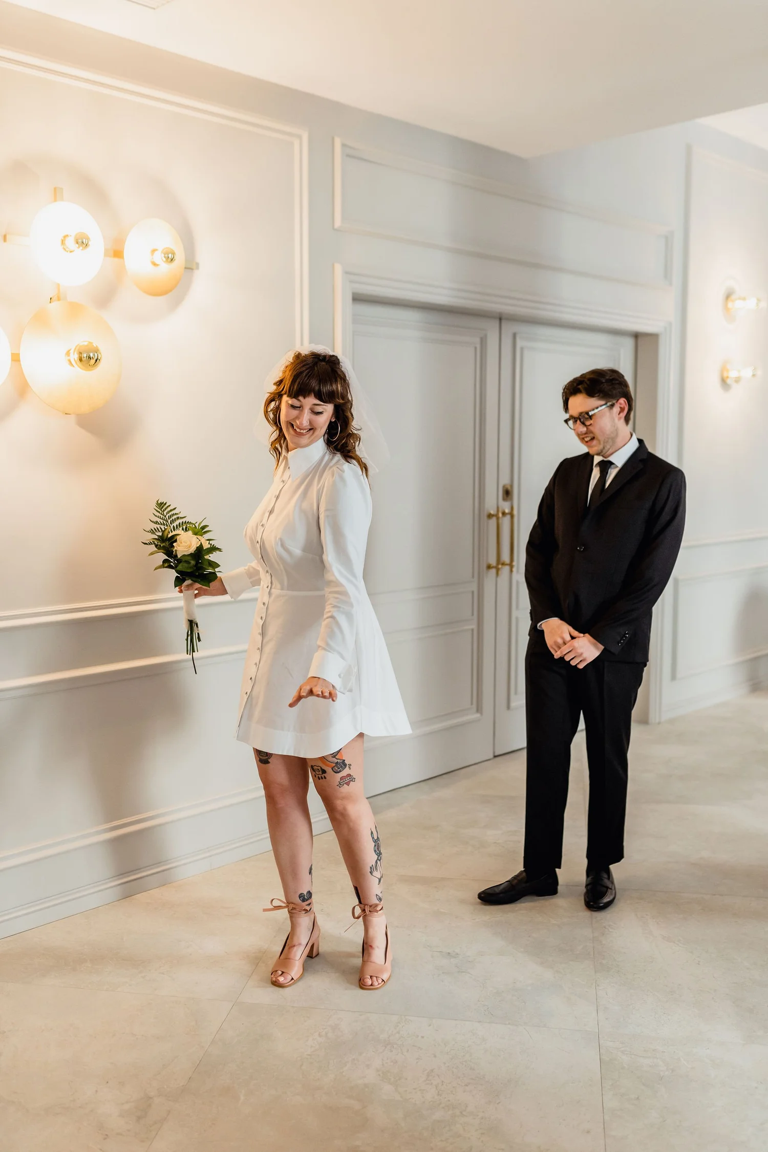 A woman in a white dress with tattoos on her legs and beige high heels holding a bouquet of white roses and greenery, is smiling and looking downward. A man in a black suit, white shirt, black tie, and glasses, stands behind her, smiling and looking