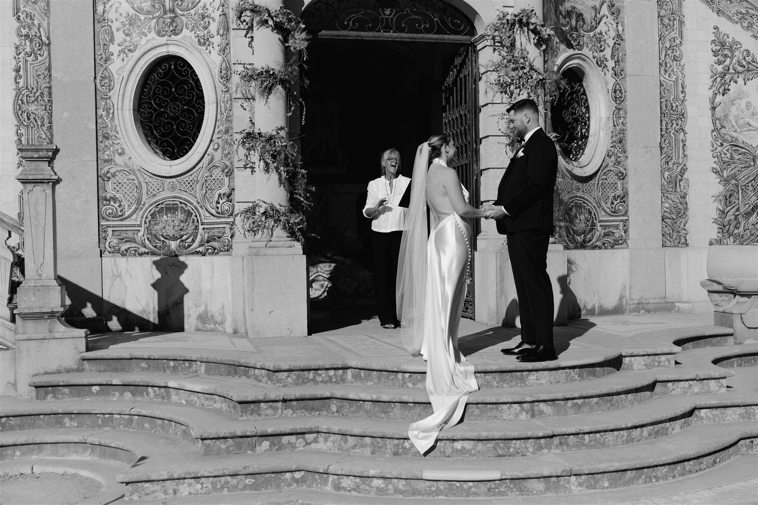 A black and white photo of a couple during a wedding ceremony on the steps of an ornate building. The bride is wearing a gown with a long train and veil, and the groom is dressed in a tuxedo. An officiant stands behind them, smiling, and they hold ha