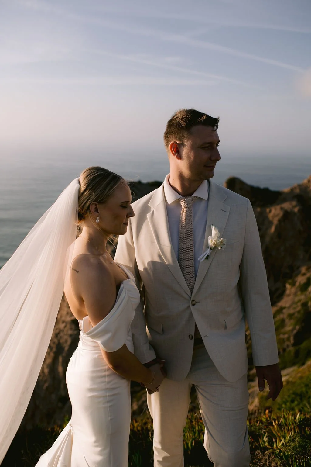 A bride and groom standing outdoors near the coast during sunset, holding hands with eyes closed or looking down, dressed in wedding attire for their elopement in Portugal.