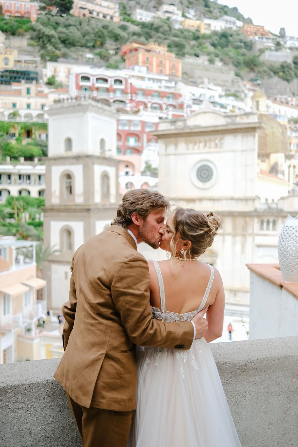 A couple in wedding attire sharing a kiss on a balcony with a hillside of colorful buildings in the background.
