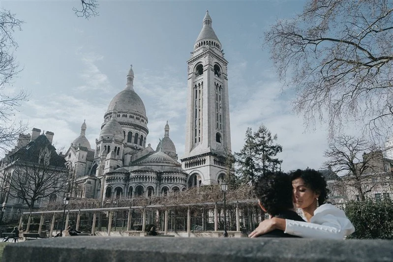Two people sitting on a park bench in front of Sacré-Cœur Basilica in Paris, France, with trees and cloudy sky in the background.