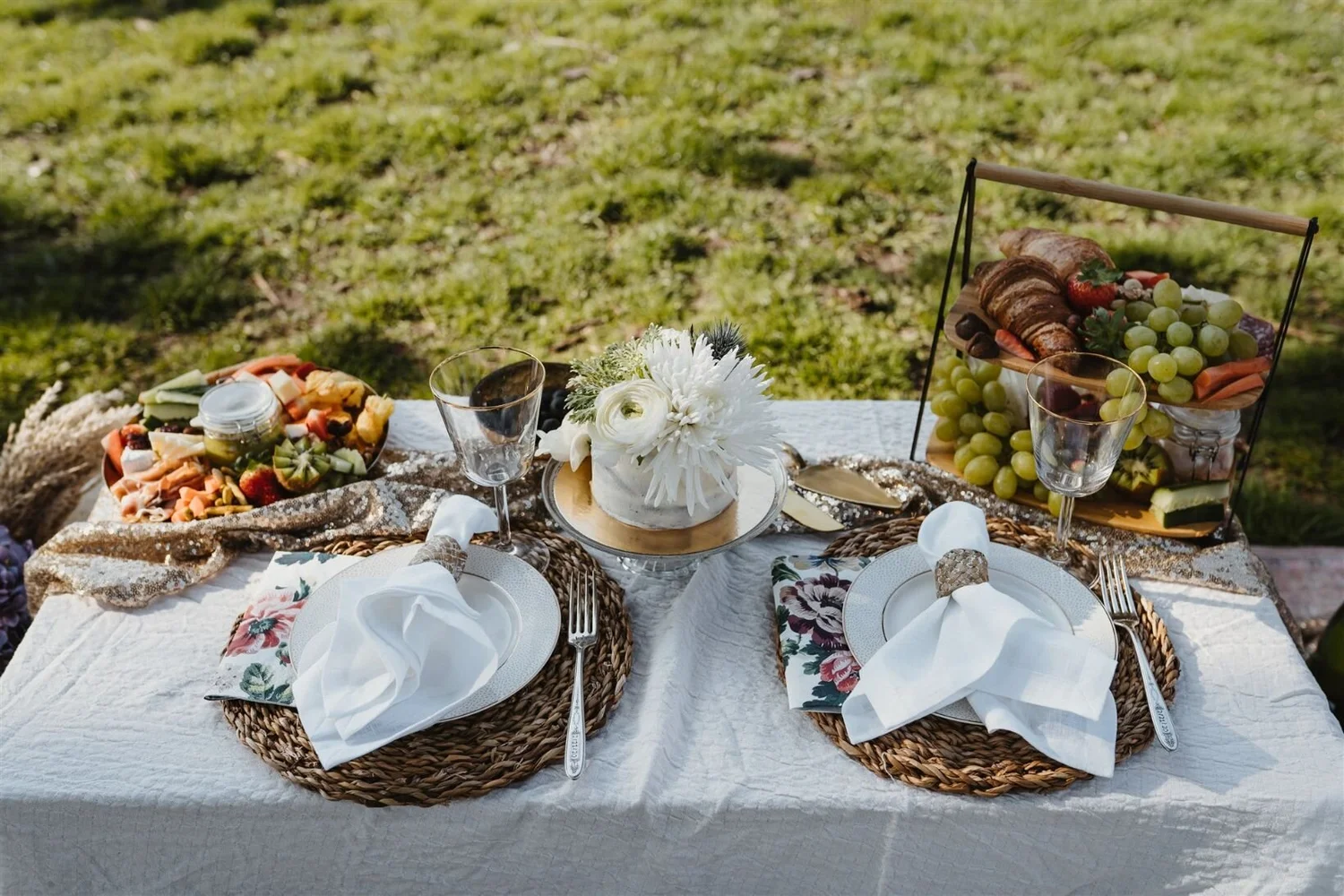 A table set for an outdoor picnic with a white tablecloth, two woven placemats, white napkins, and silverware. In the center, there's a cake with white flowers. To the left, a bowl of assorted fruit and cheese, and to the right, a tiered wooden tray