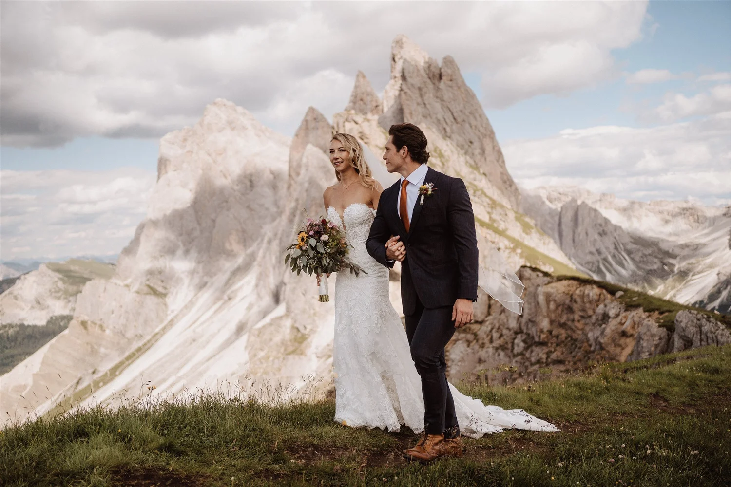 A bride and groom walking together outdoors in a mountainous landscape. The bride wears a white lace wedding dress and holds a bouquet, while the groom is dressed in a dark suit with a white shirt and an orange tie. The sky is partly cloudy.