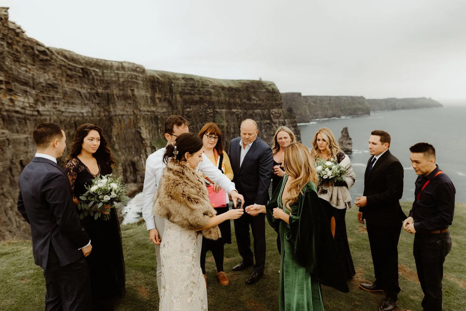 A group of people dressed in formal and traditional attire, standing outdoors on a grassy area near the cliffs by the ocean, participating in a wedding ceremony.