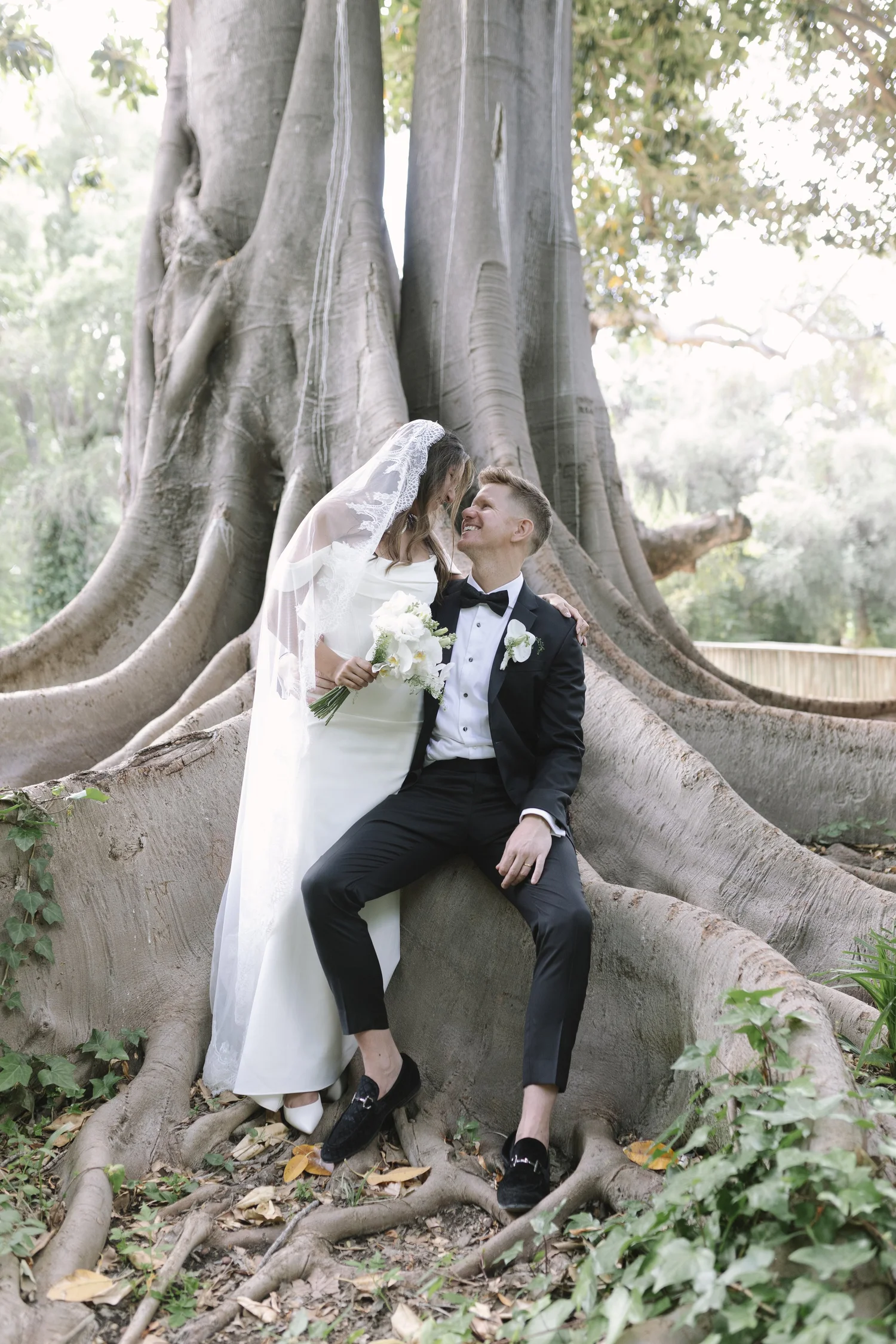 A bride and groom smiling at each other outdoors near a large tree with exposed roots.