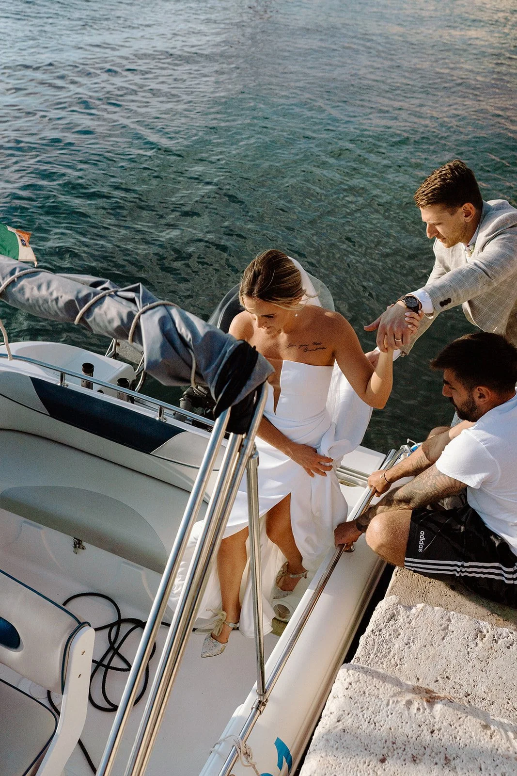 A woman in a white dress stepping onto a boat from a pier, assisted by a man in a light-colored suit, with another man nearby. The scene takes place on a body of water during sunset.