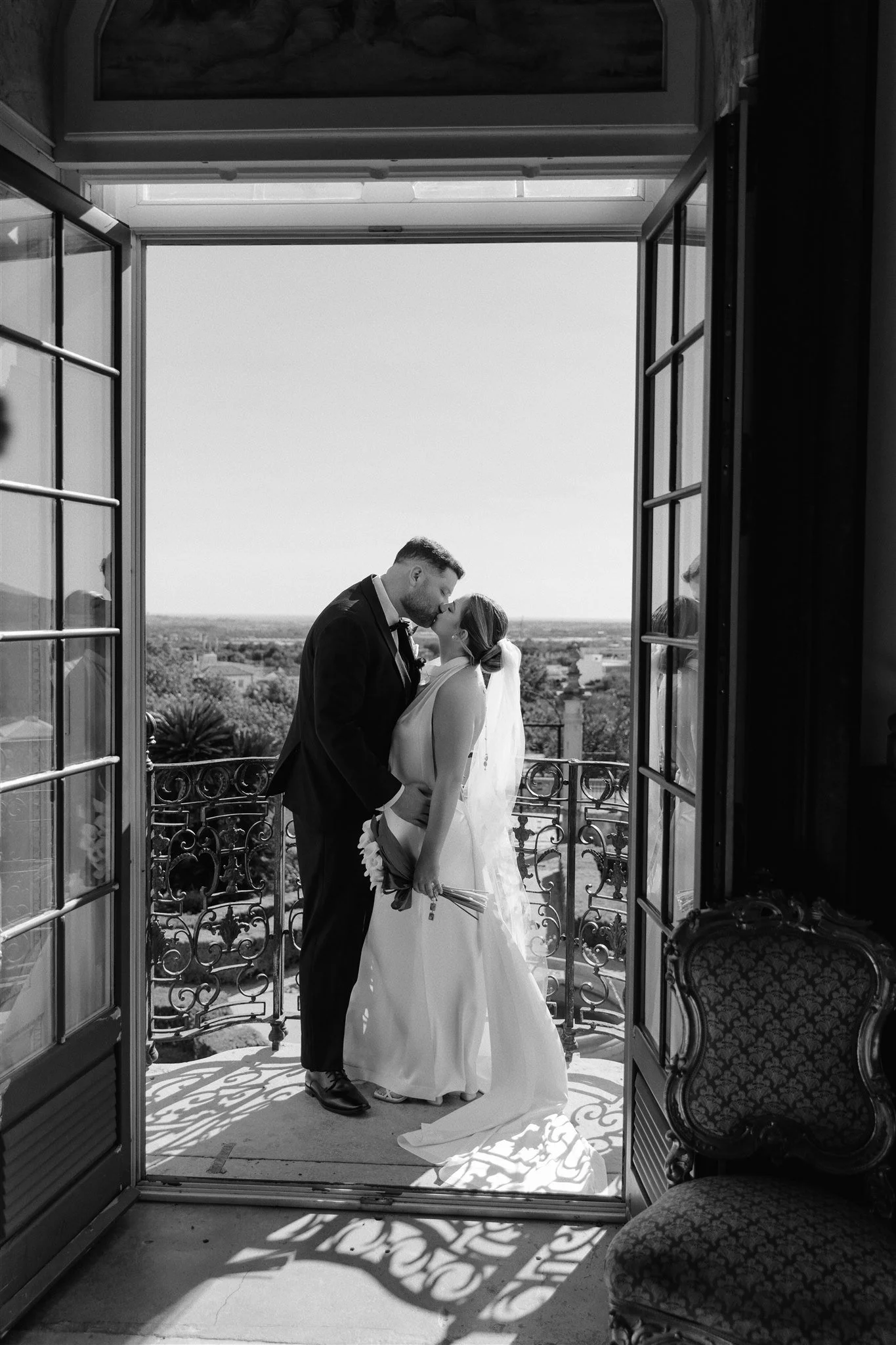 A black and white photo of a bride and groom kissing on a balcony with open French doors. The groom is wearing a tuxedo and the bride is in a wedding dress holding a bouquet. Shadows cast intricate patterns on the floor from the balcony railing, and