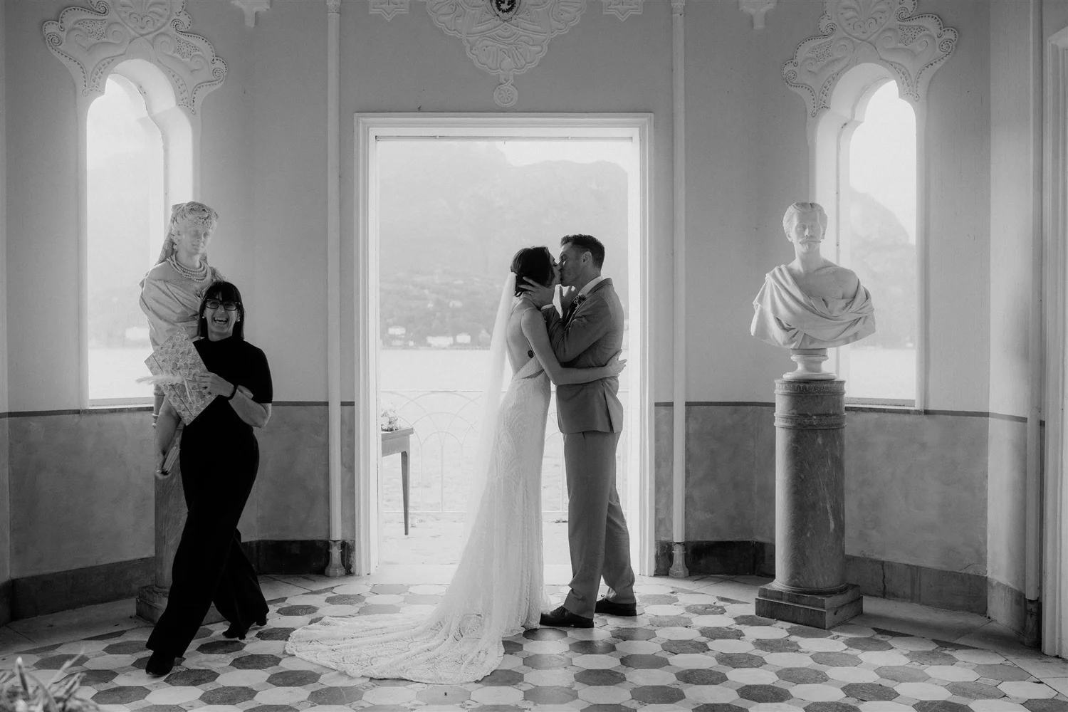A black and white photo of a bride and groom sharing a kiss in a decorated indoor space with large windows and classical sculptures. A woman in glasses holding a bouquet is smiling on the left, while a woman in earrings and a patterned dress is smili