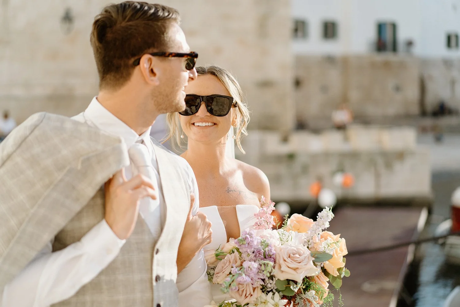 A smiling bride holding a bouquet of flowers, standing next to a groom with a jacket draped over his shoulder, both wearing sunglasses, in an outdoor setting by the water.