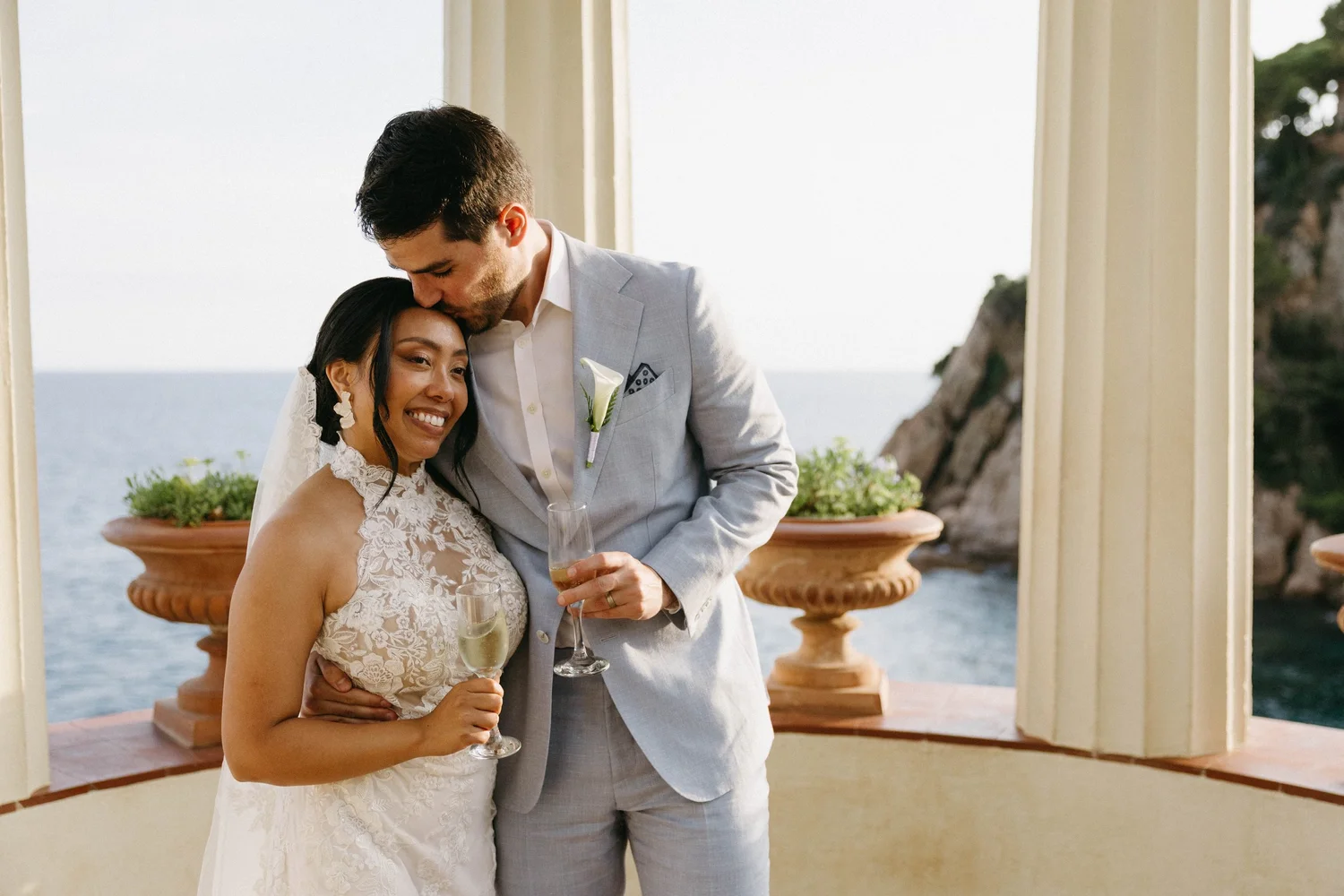 A man and woman in wedding attire sharing a tender moment with glasses of champagne, overlooking the ocean from a terrace with large planters.