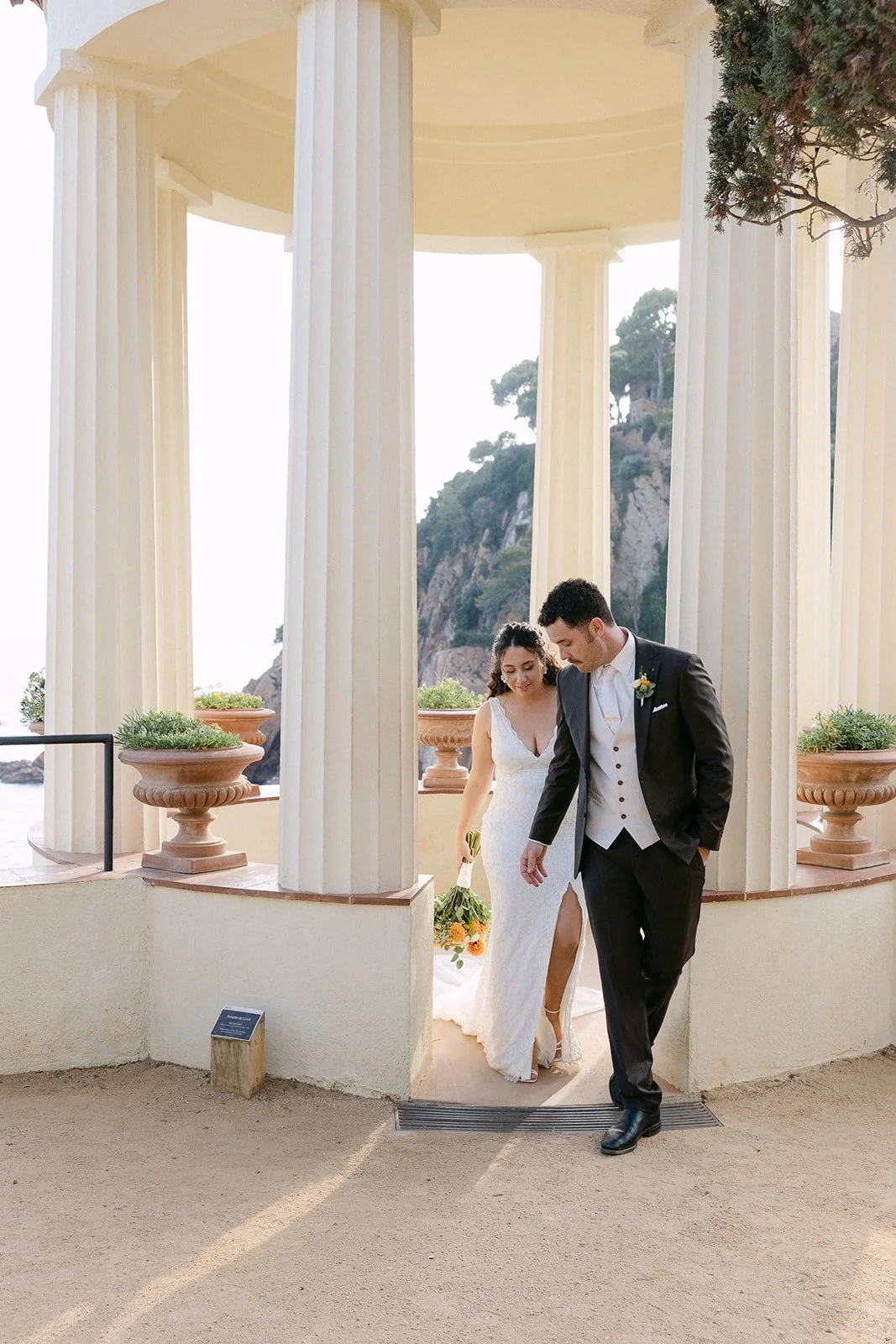 A bride and groom walking together beneath a classical pavilion with white columns, with a scenic rocky hillside in the background.