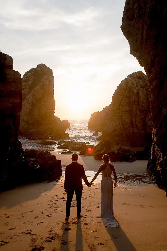 A couple in wedding attire holding hands and walking on a sandy beach between large rocky cliffs at sunset, with the ocean in the background.