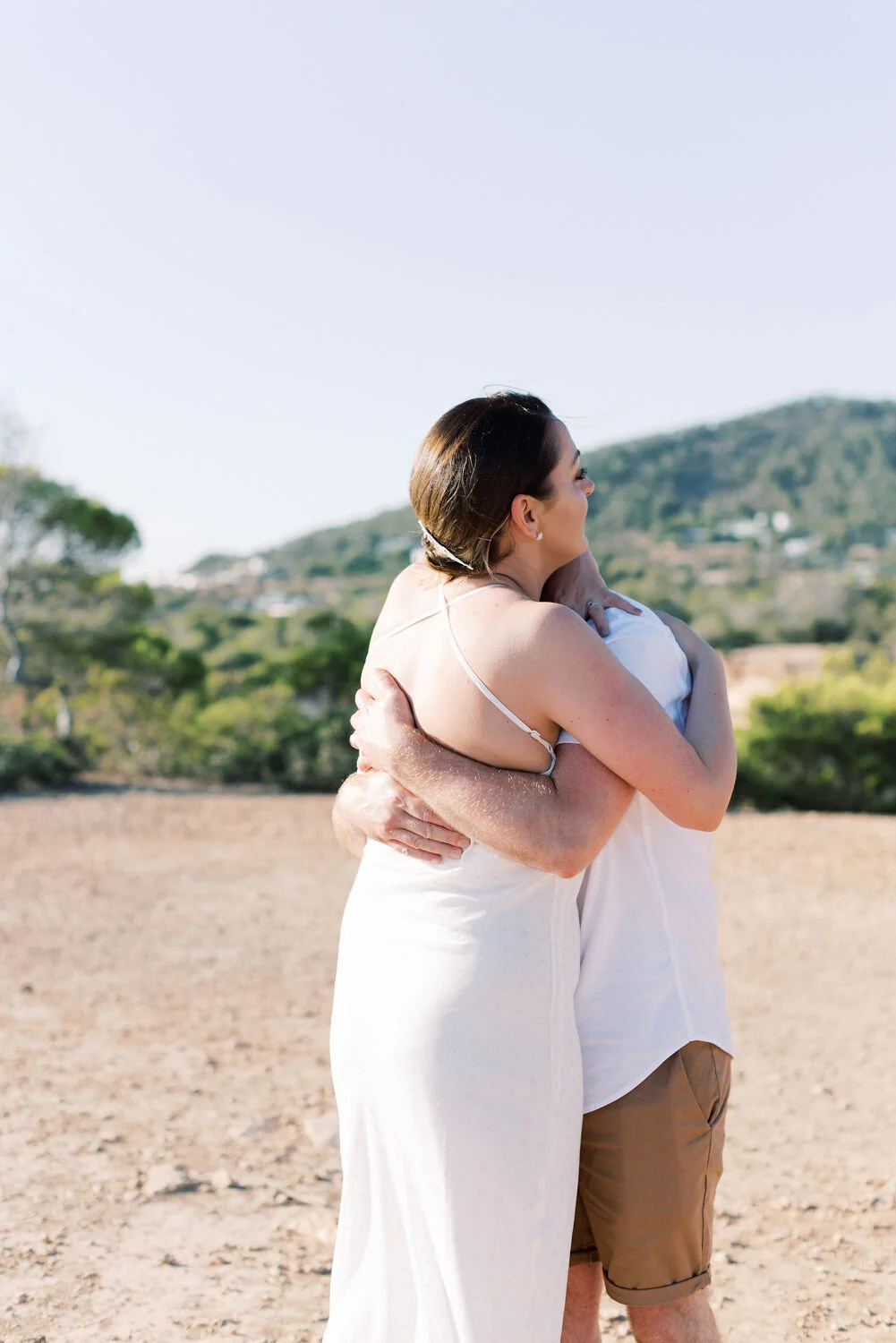 A woman in a white dress hugging a person in a white shirt outdoors with hills and trees in the background.