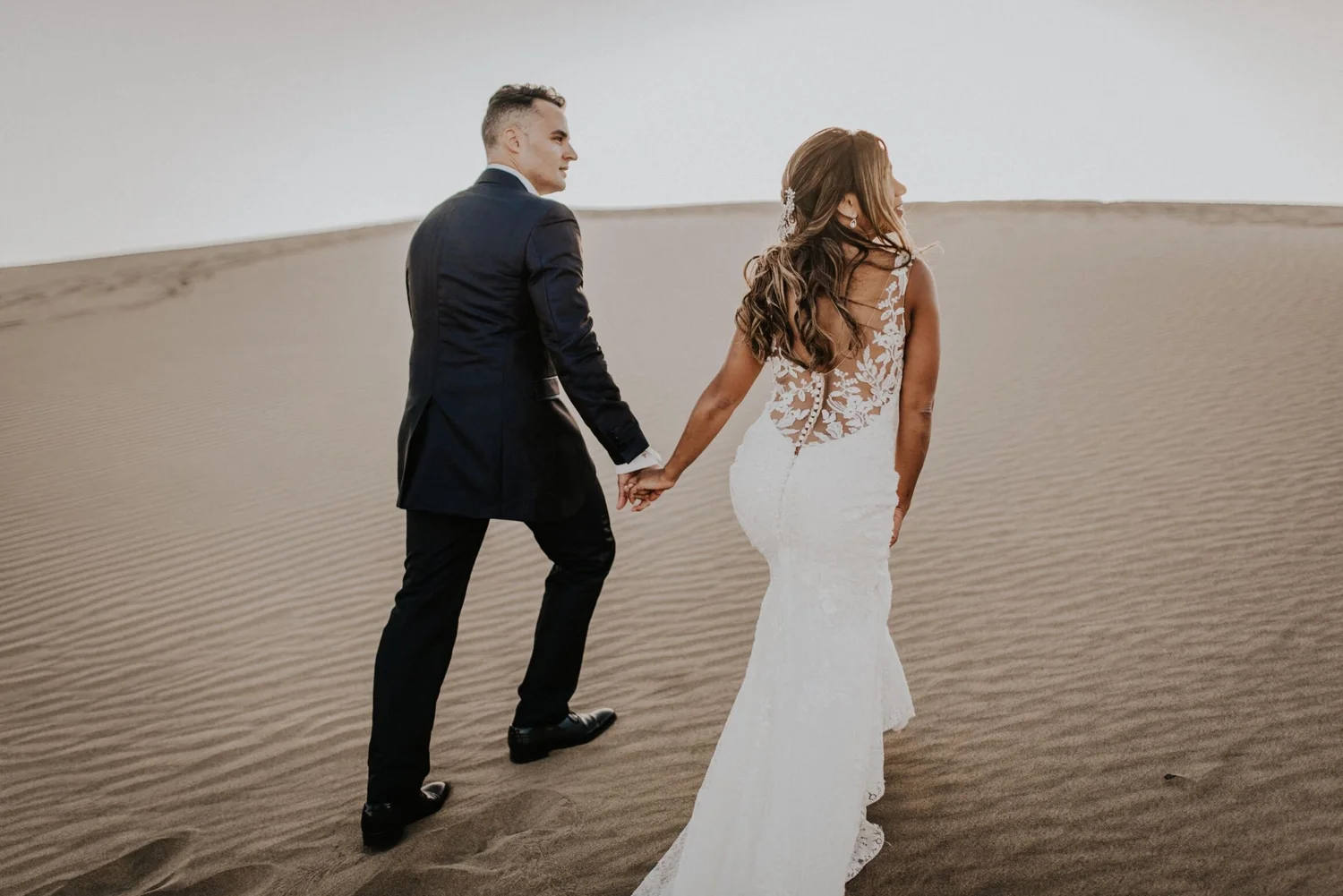 A bride and groom holding hands and walking in a desert with a white silhouette cutout of the bride in front of them.