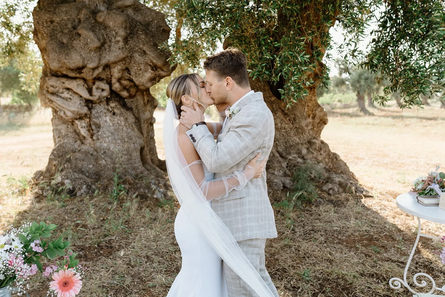 A bride and groom kiss outdoors under a large tree, with flowers and wedding decorations nearby.