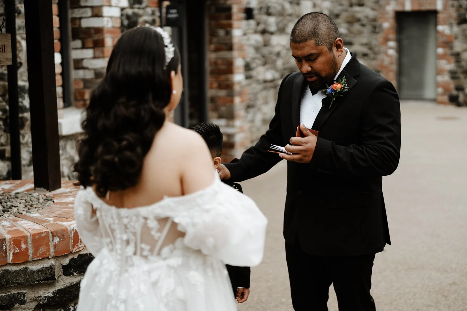 A wedding ceremony with a man in a black tuxedo and a woman in a white off-the-shoulder lace wedding dress, standing outside near a brick wall. The man appears to be reading vows or a speech while the woman listens. The scene is intimate and formal.