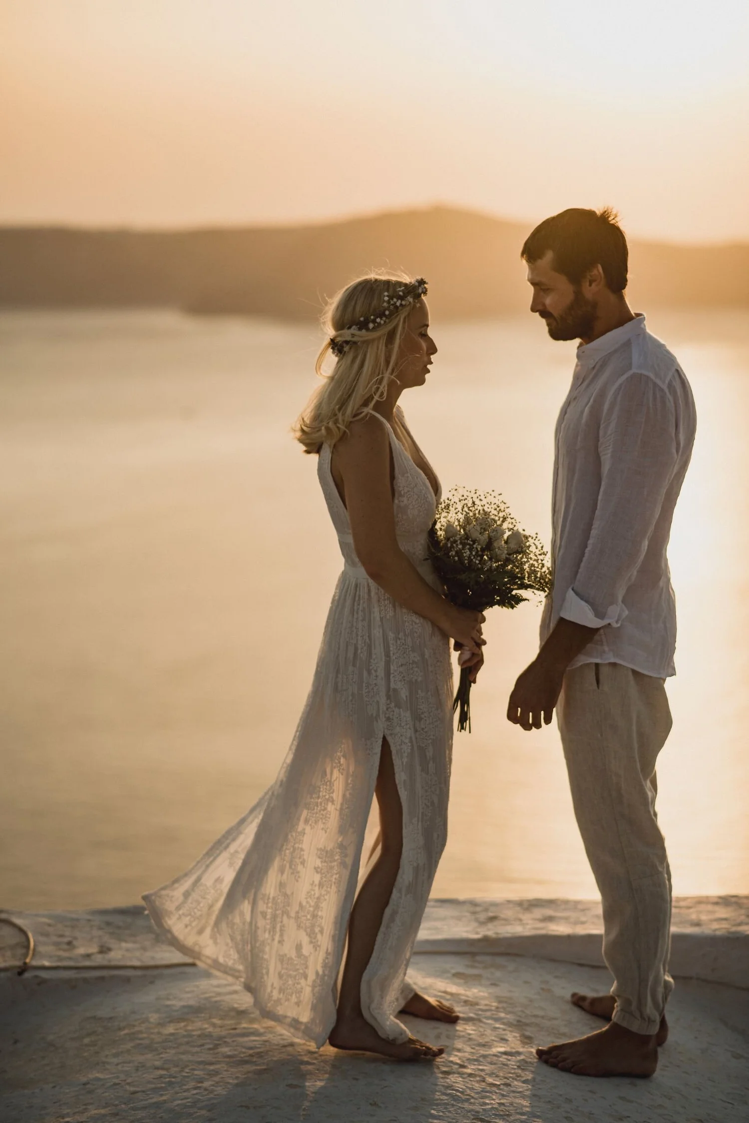 A couple stands close together on a beach at sunset, holding hands and gazing at each other, with a mountain in the background.