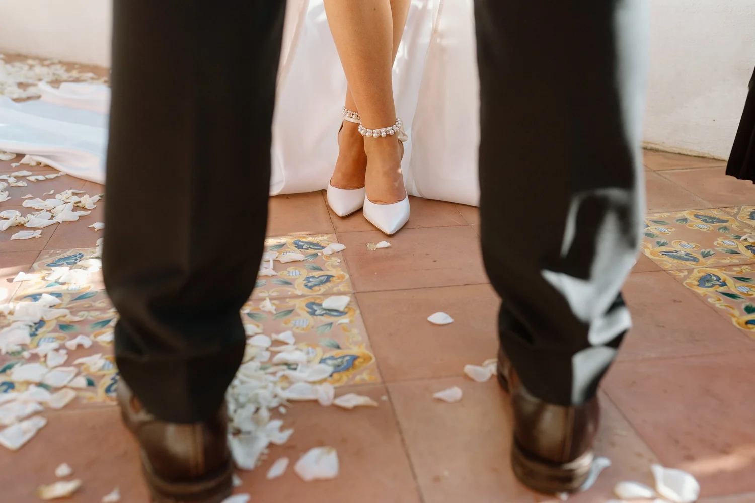 View of a bride's legs wearing pearl bracelet and white heels, seen through the legs of two men in black pants, during a wedding ceremony with flower petals scattered on the floor.