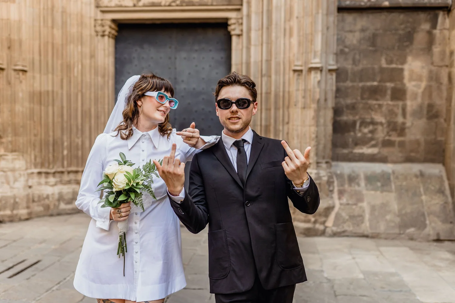 A couple dressed in wedding attire, wearing sunglasses, standing outdoors in front of an old stone building. The woman holds a bouquet of white flowers and greenery and is smiling, while the man makes a gesture with both hands, smiling slightly.