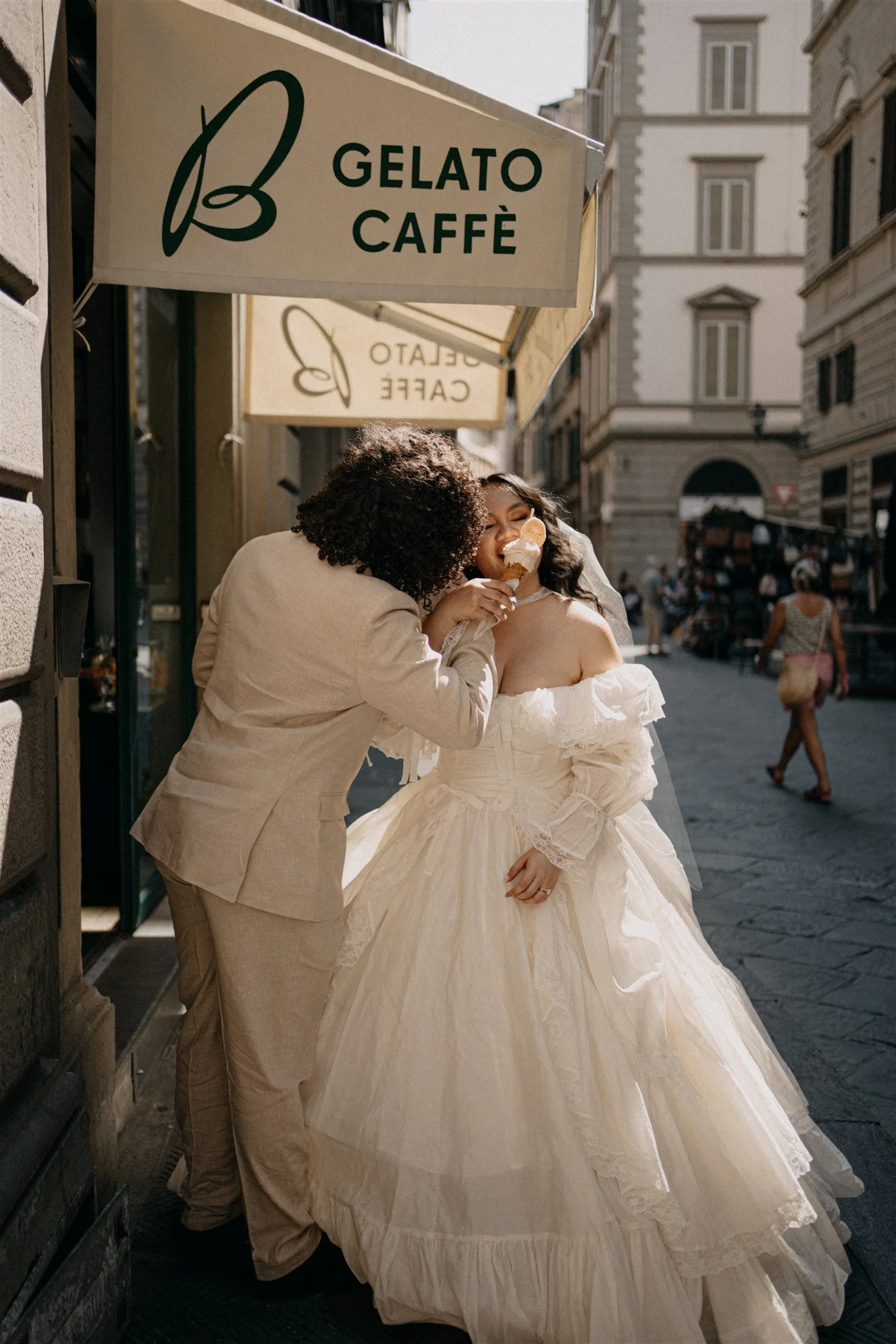 A bride in a white wedding gown is being kissed by a person in a tan suit on a city street. The bride is holding an ice cream cone and smiling with her eyes closed. There are buildings and pedestrians in the background, and signs above them read "Gelato Caffè".