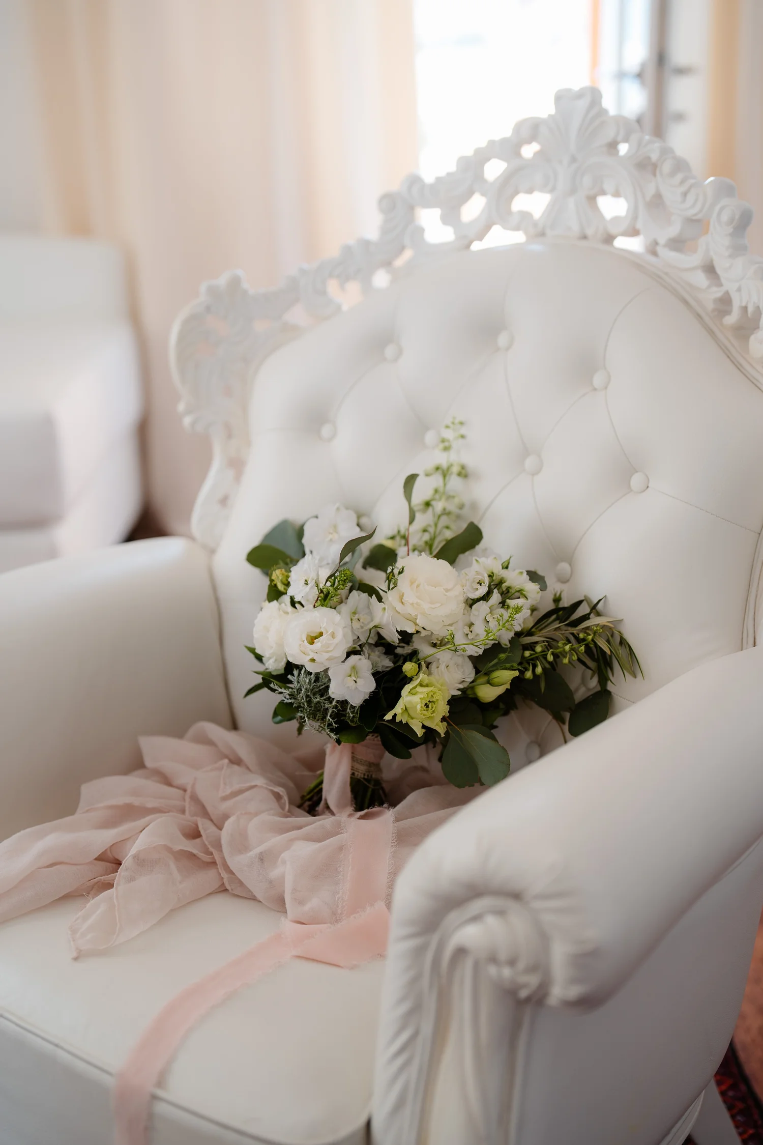 A white ornate vintage-style armchair with a tufted backrest and carved decorative details, decorated with a bouquet of white flowers and greenery, and soft pink fabric draped on it.