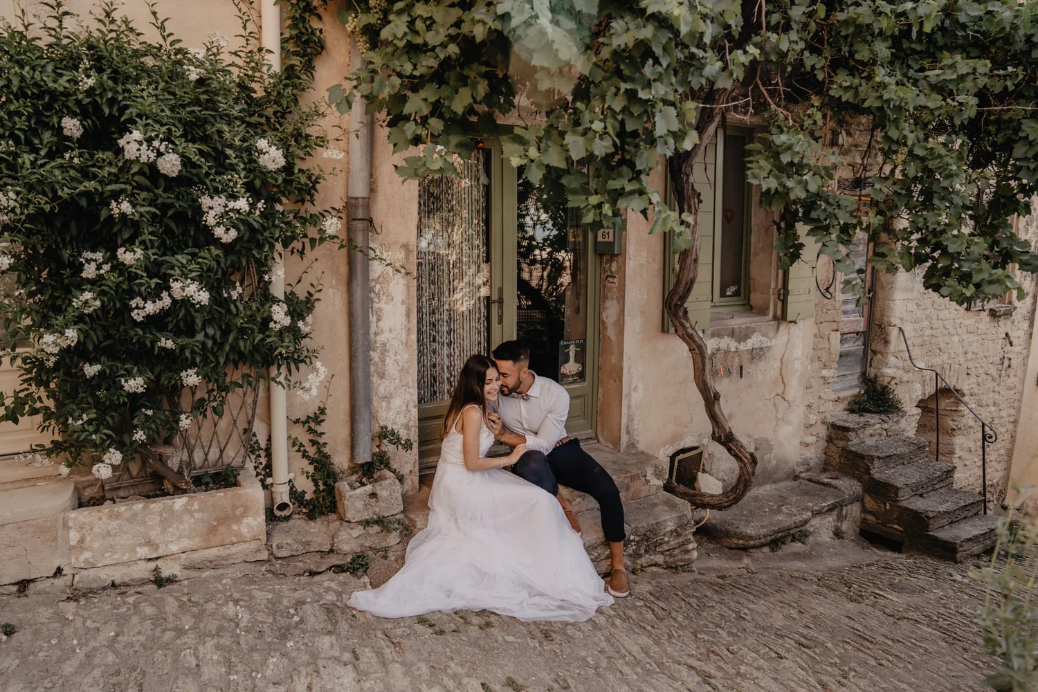 A couple sitting on stone steps outside a rustic building, with vines and flowers overhead, the woman in a white dress and the man in a white shirt and bow tie, sharing a moment.