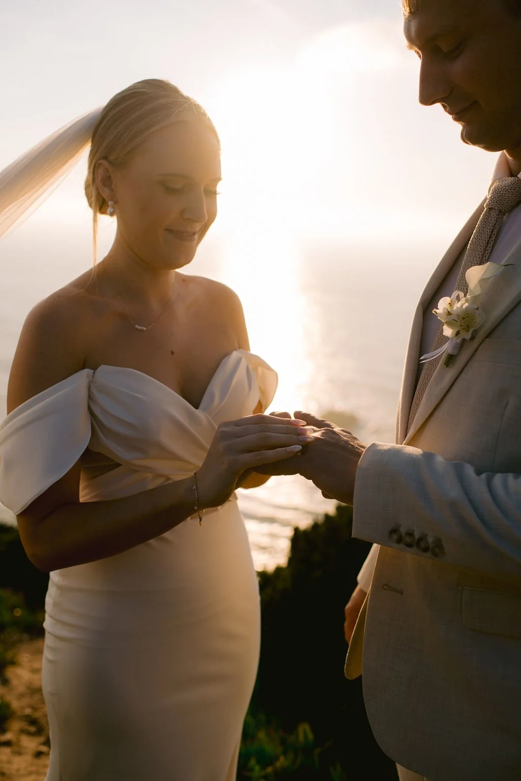 A bride and groom during a wedding ceremony on the beach at sunset, holding hands and smiling gently at each other.