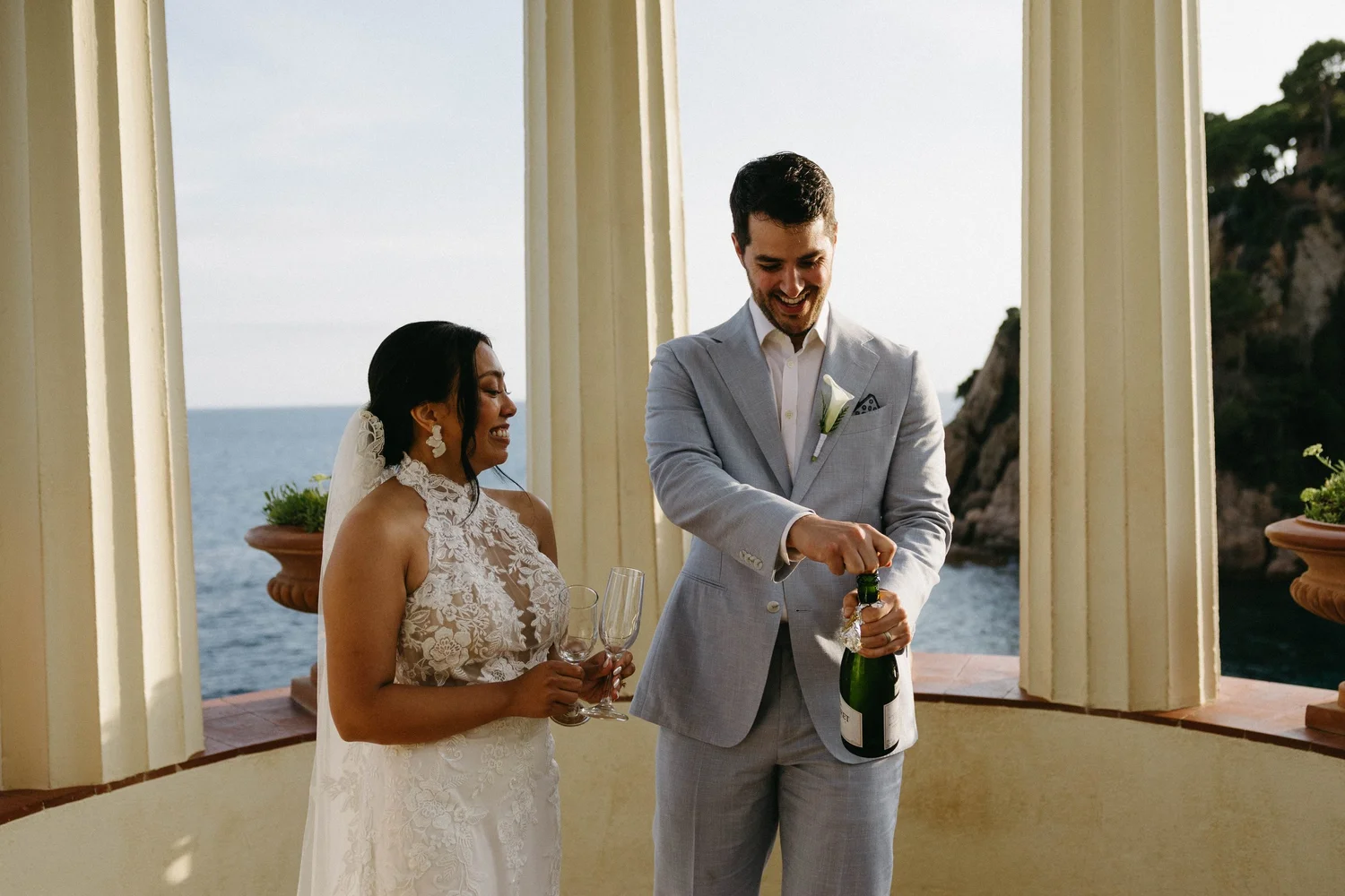 A bride and groom standing under a pavilion with the ocean in the background, celebrating their wedding with champagne, on a sunny day.