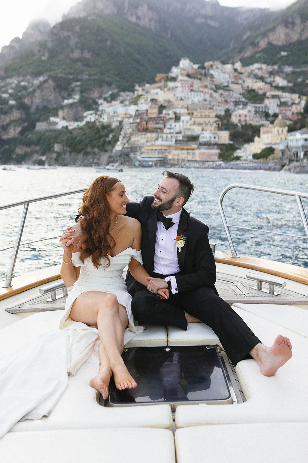 A newlywed couple sitting closely on a yacht, enjoying each other's company, with a coastal town and mountain in the background.