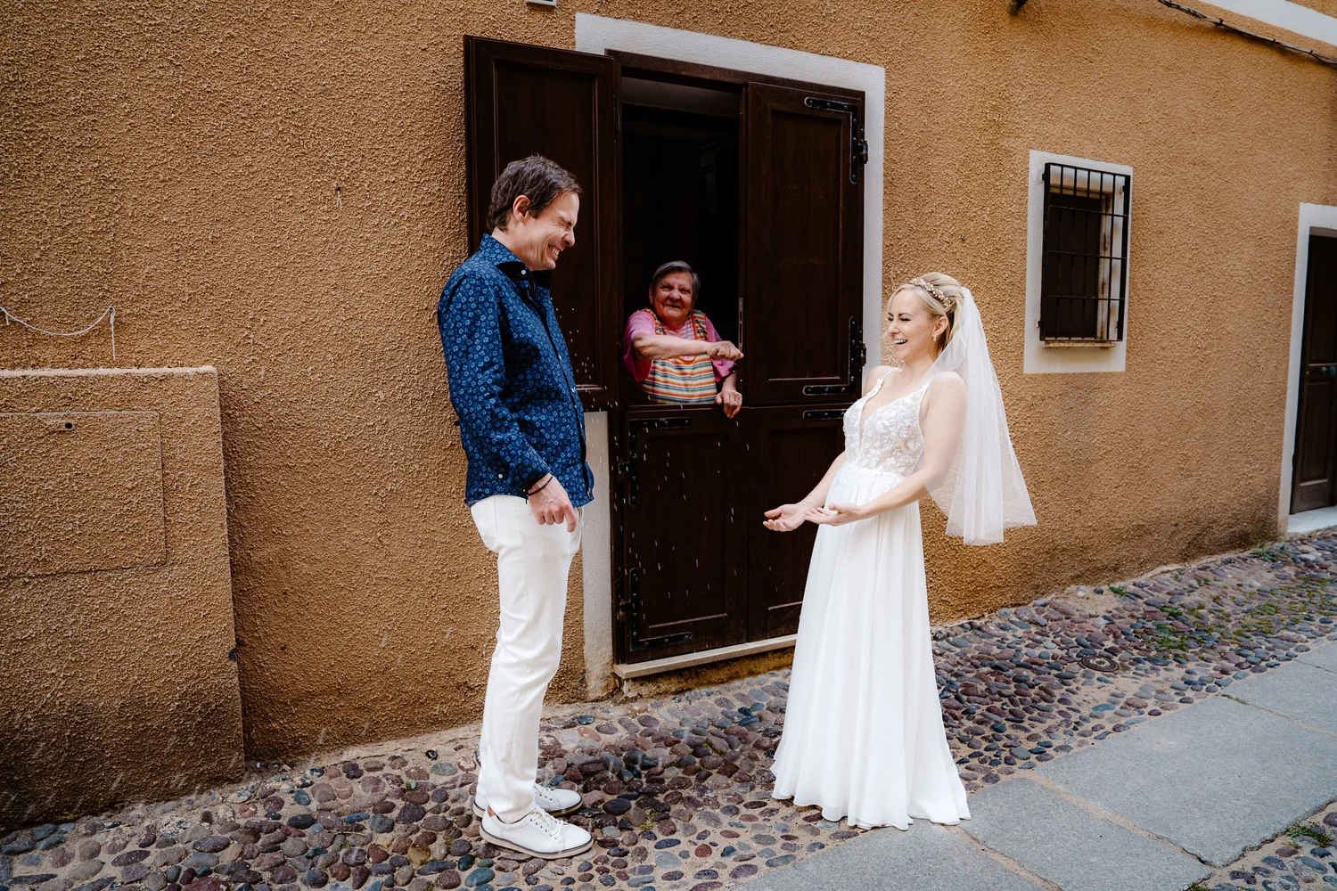 A bride in a white wedding dress and veil, smiling with a man in a blue shirt and white pants, outside near a brown wall, with an elderly woman smiling from a window.