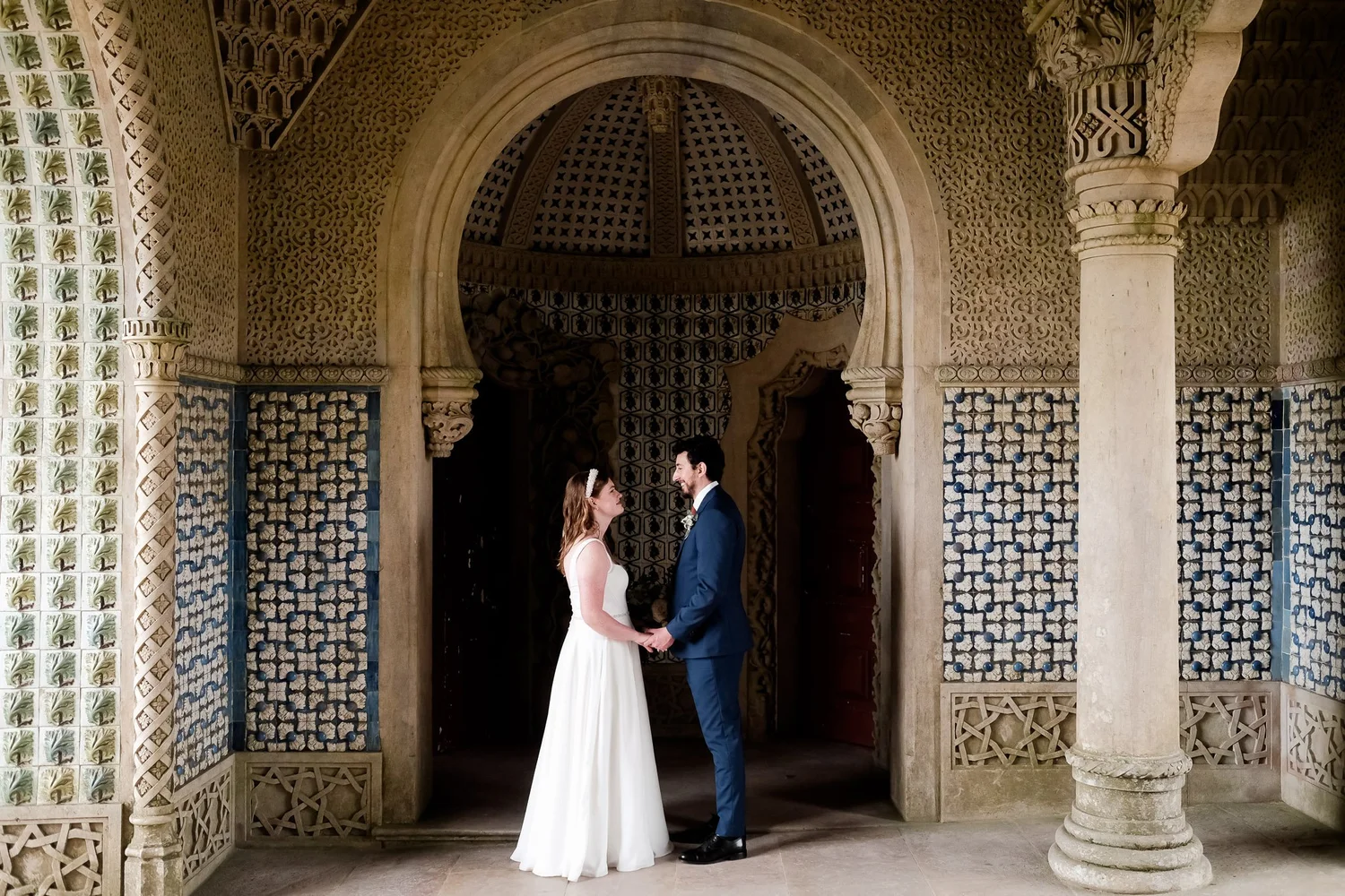 A bride and groom standing face-to-face under an ornate stone archway, holding hands, with intricate architectural details and patterned walls in the background.