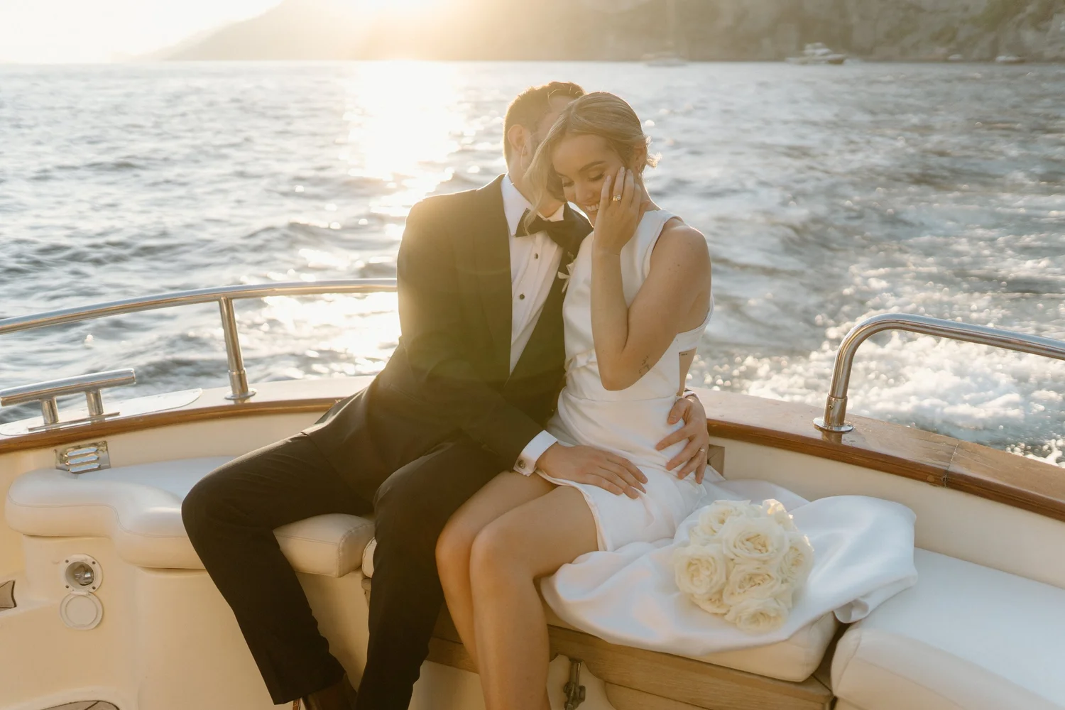 A newlywed couple dressed in wedding attire sitting on a yacht with water and shoreline in the background, sharing a tender moment at sunset.
