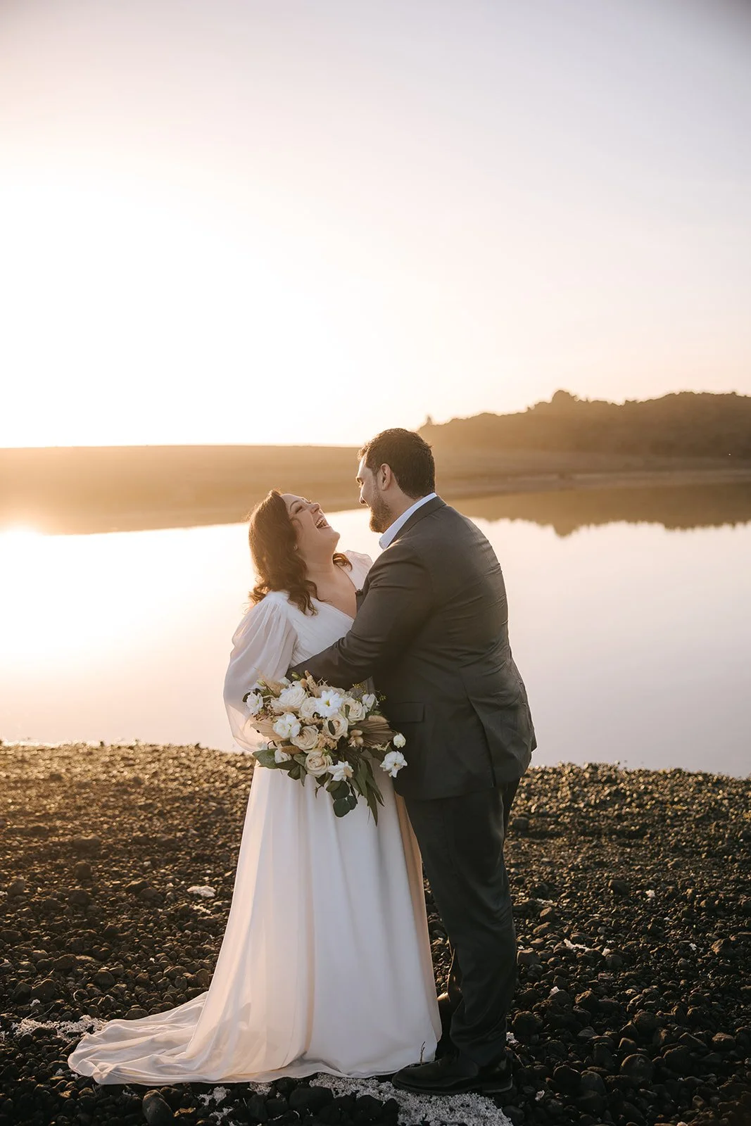 A bride and groom smiling and embracing by a lake at sunset. The bride is holding a bouquet of white flowers and wearing a white dress, the groom is dressed in a dark suit.