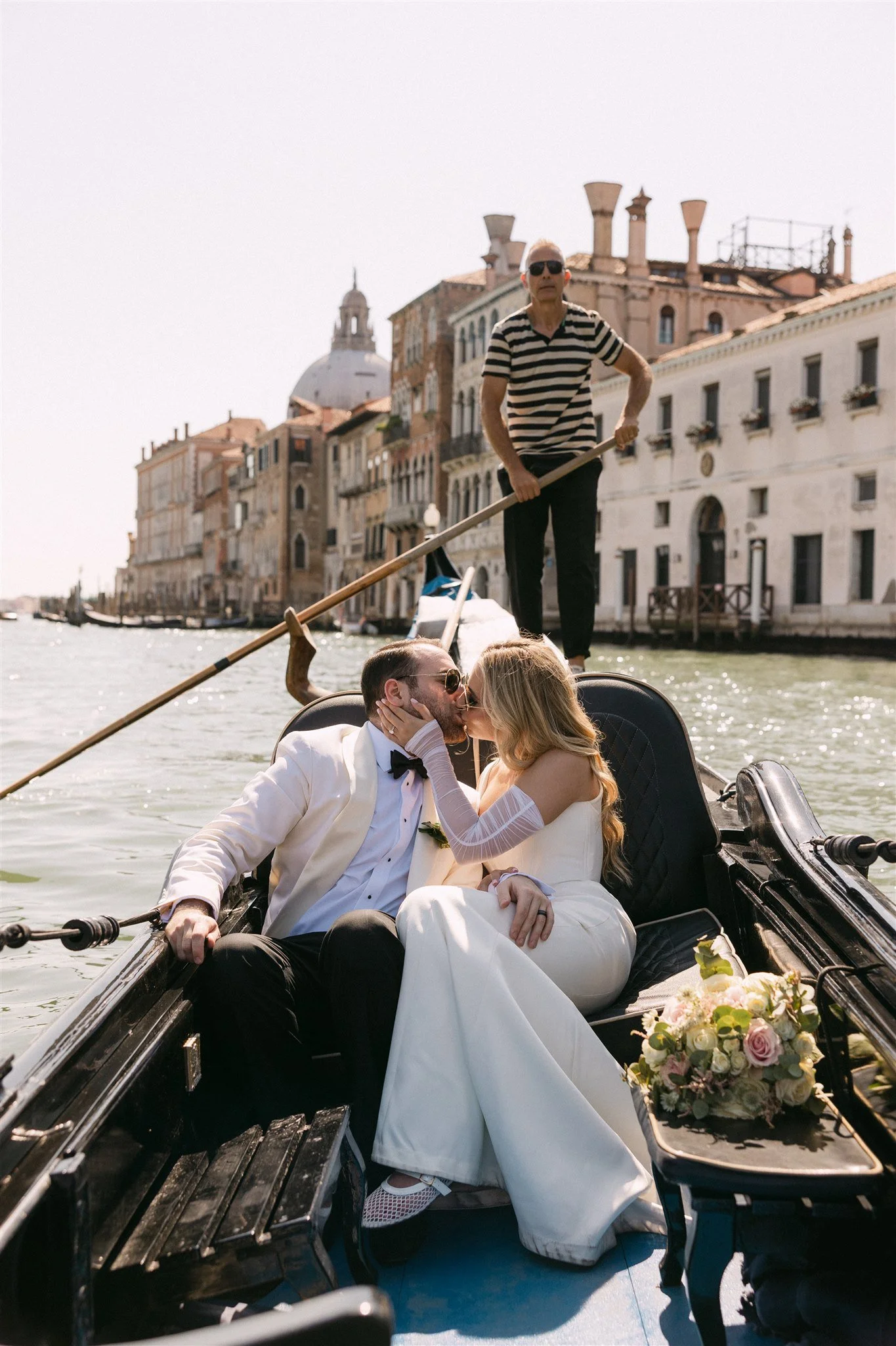 A newly married couple kissing on a gondola ride in Venice, Italy, with a gondolier standing behind them, wearing sunglasses and a striped shirt, with historic buildings and a domed church in the background.