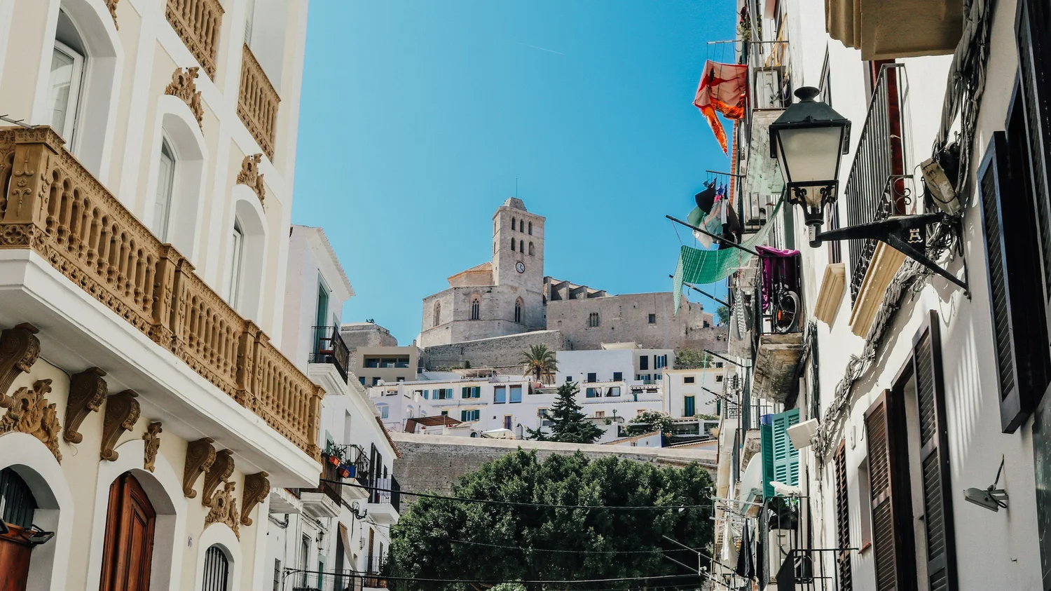 Narrow street with ornate white buildings on the left and balconies on the right, laundry hanging from lines, and a historic stone tower atop a hill in the background under a blue sky.