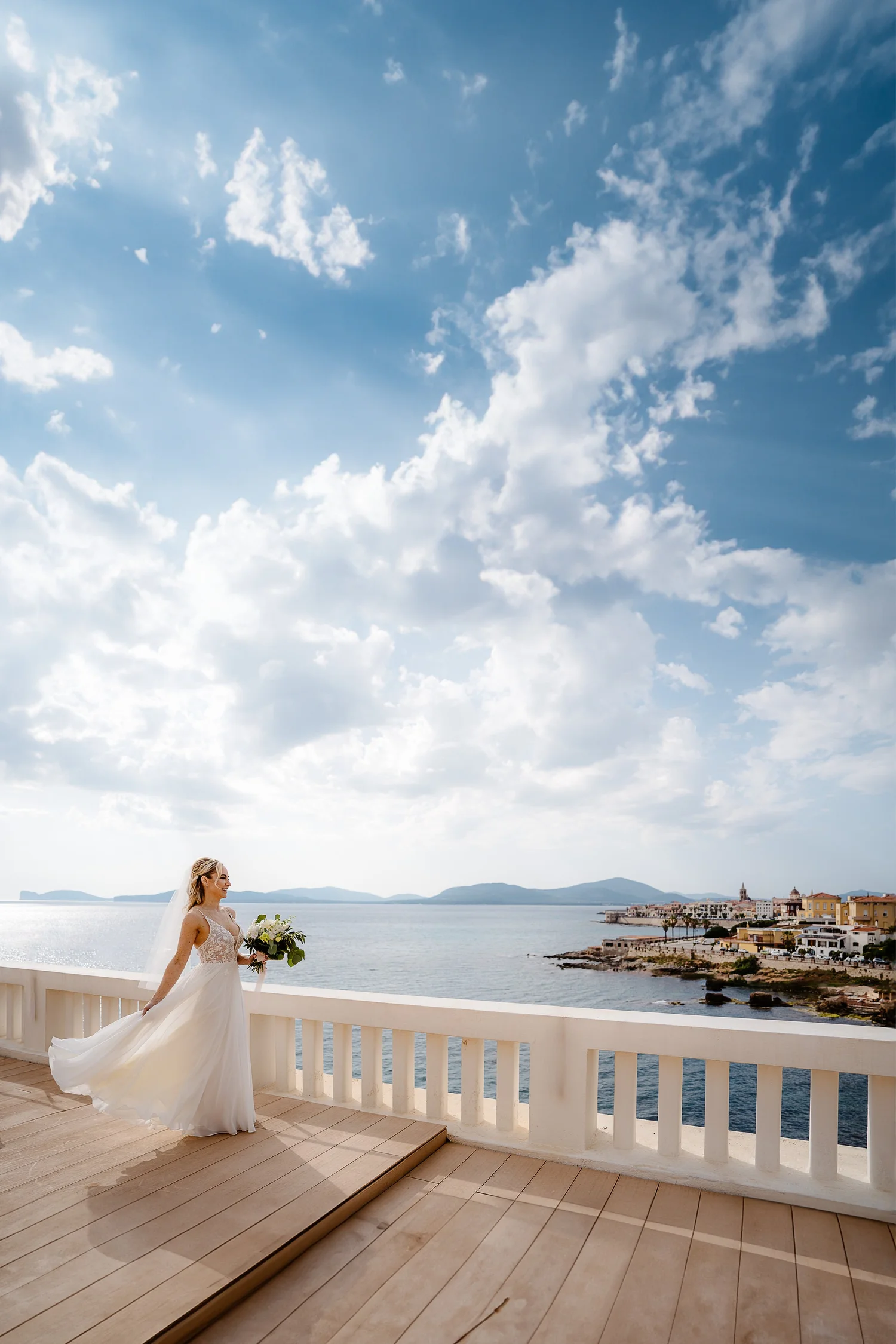 A bride in a white wedding dress holding a bouquet of flowers, standing on a balcony overlooking a coastal town and ocean under a partly cloudy sky.
