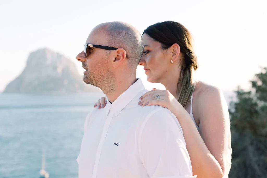 A couple on a boat, with the woman hugging the man's shoulders from behind near a body of water, with a large rock formation in the background.