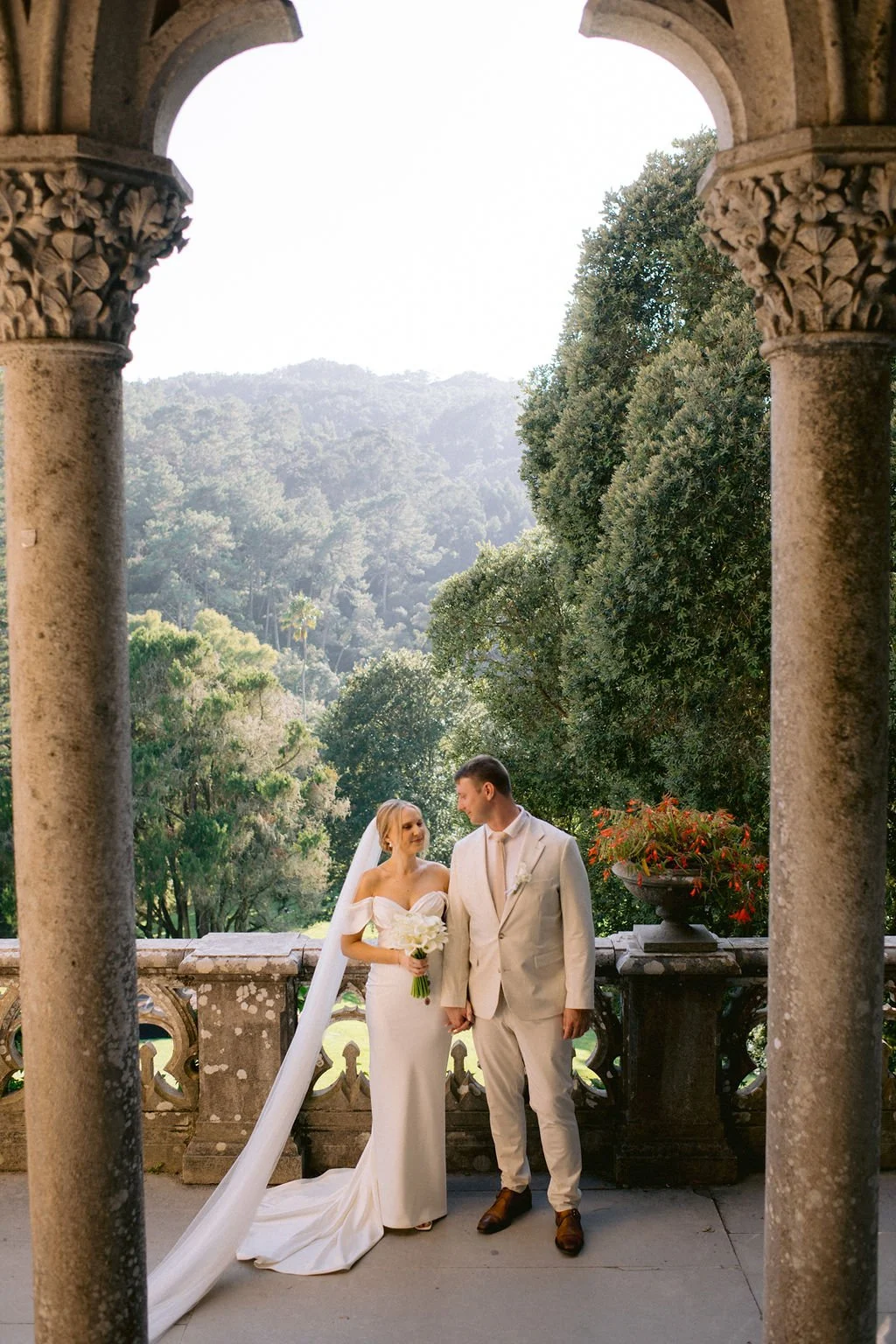 A bride and groom holding hands, standing on a stone balcony with ornate columns, overlooking a lush green landscape and mountains.